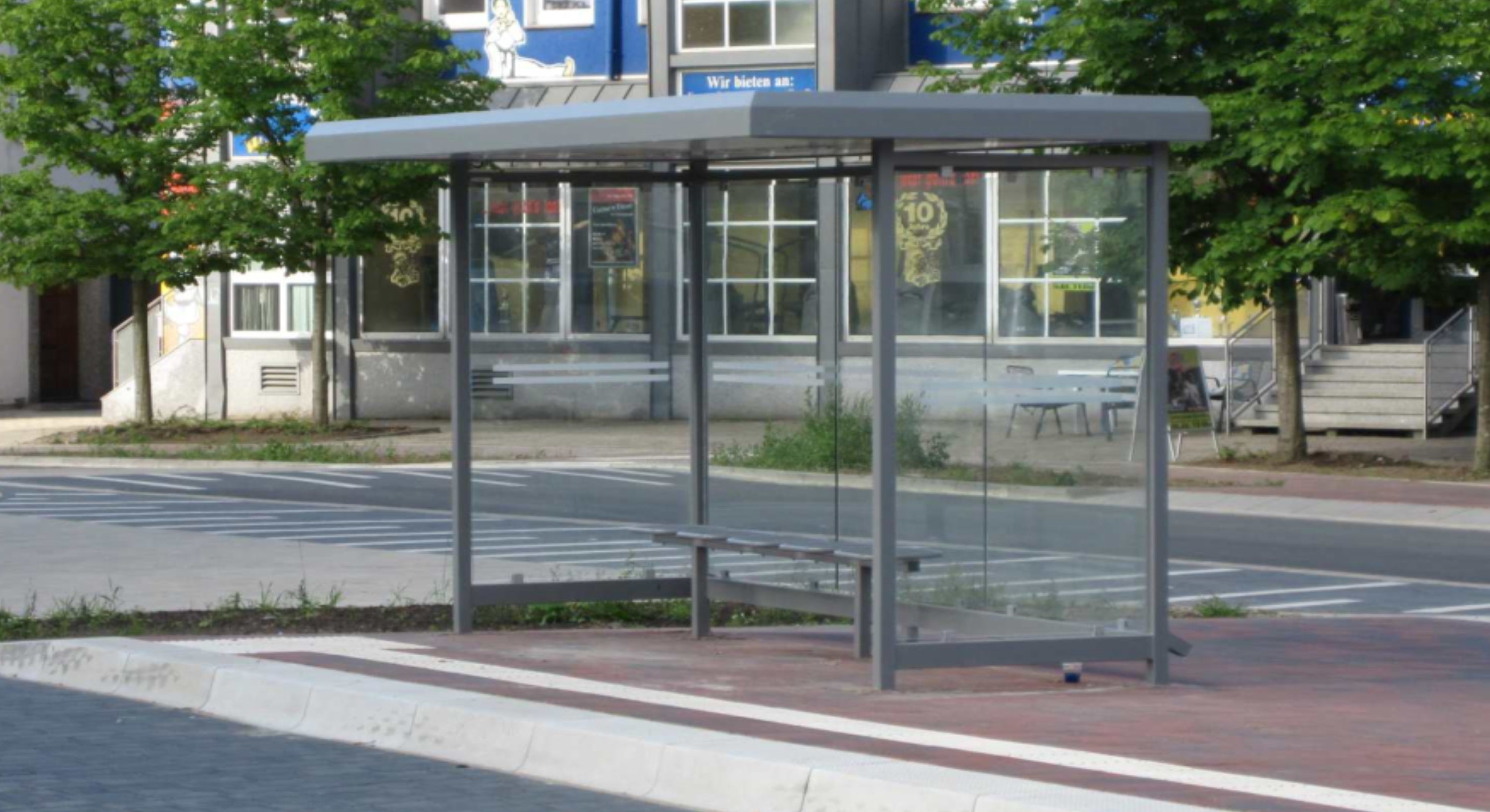 Ein moderner, gläserner Fahrgastunterstand mit Sitzbank und Überdachung, umgeben von städtischer Umgebung und Bäumen.