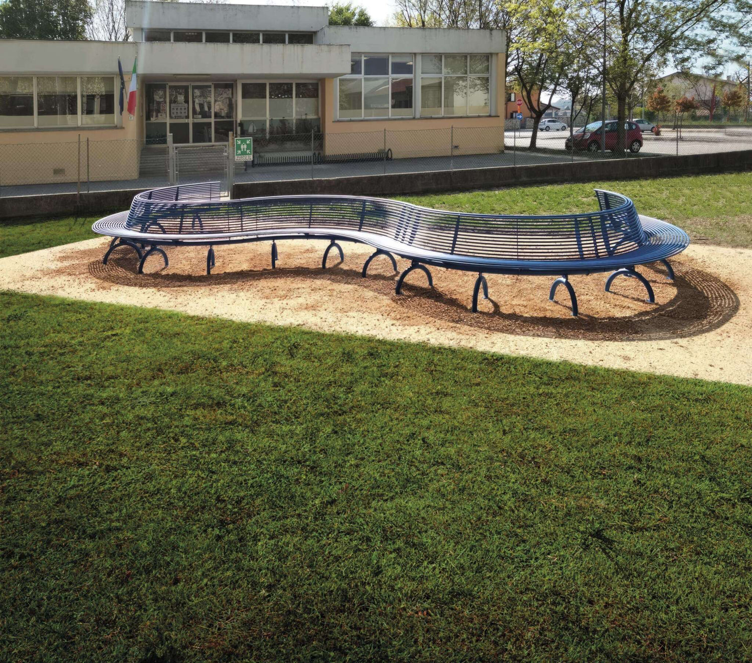 Auf dem Bild ist eine große, geschwungene blaue Metallbank zu sehen, die sich auf einer kreisförmigen Sandfläche befindet. Die Bank steht im Freien in einem Park oder Schulhof, umgeben von grünem Rasen. Im Hintergrund erkennt man ein Gebäude mit großen Fenstern und zwei Flaggen, wahrscheinlich eine Schule oder öffentliche Einrichtung. Es gibt einen Zaun, einige Bäume und geparkte Autos neben einer asphaltierten Fläche. Die Szene wirkt ruhig und gepflegt.