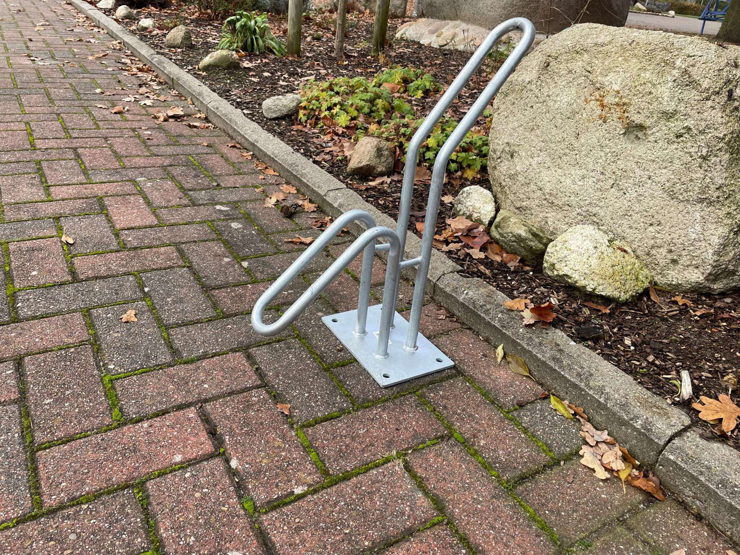 Silberner Fahrradständer auf gepflastertem Weg neben Steinen und Pflanzen im Freien.