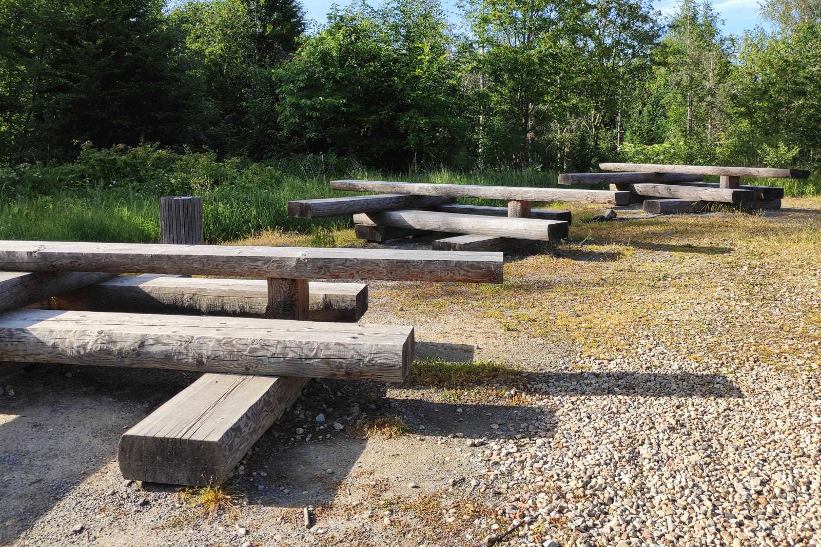 Holzbänke in einem parkähnlichen Bereich, umgeben von Bäumen und Kieselsteinen.