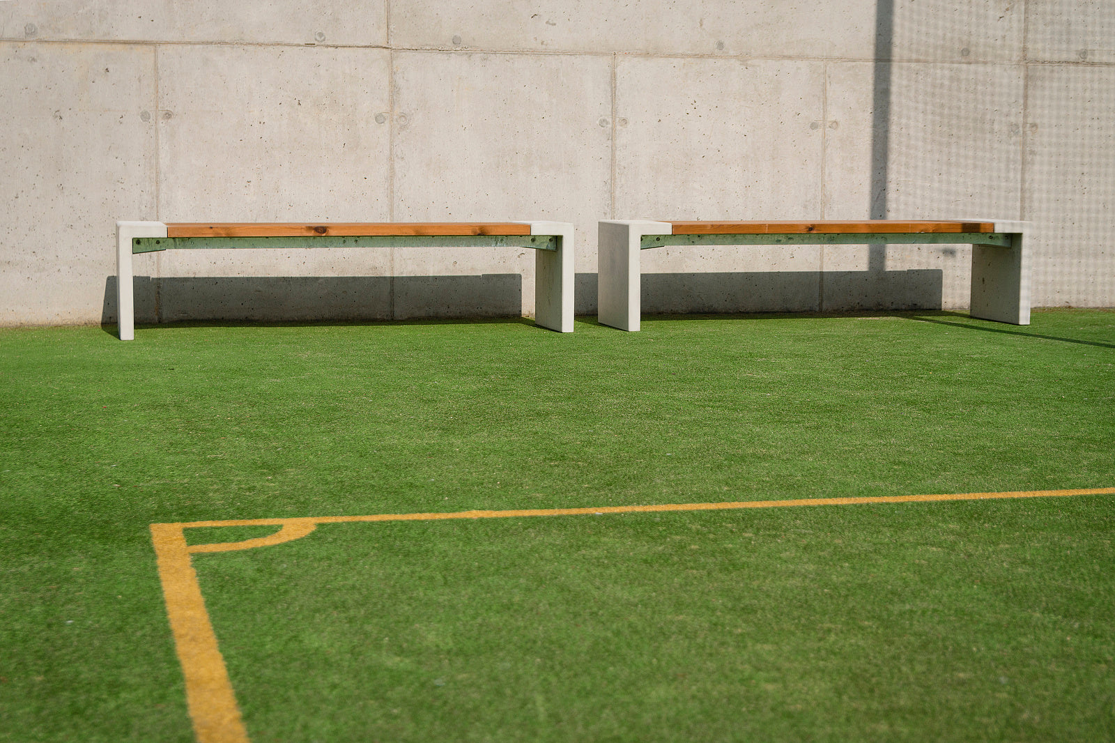 Zwei moderne Bänke auf Kunstrasen vor einer Betonwand, ideal für Außenbereiche und Sportplätze.