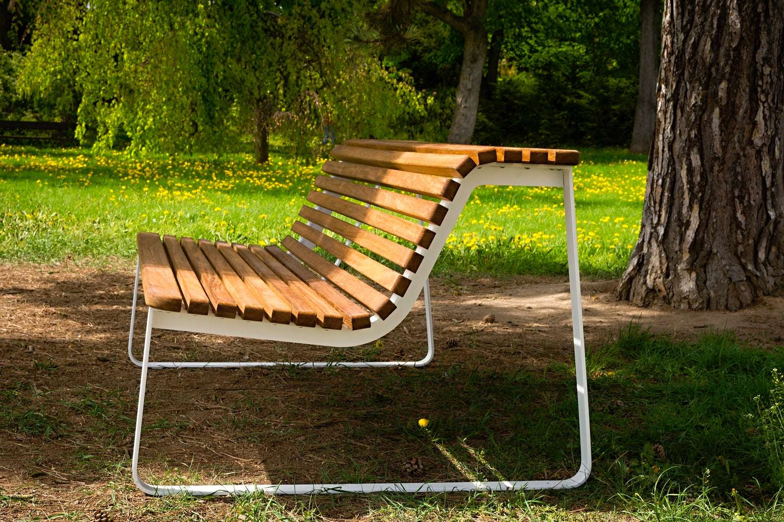 Moderne Parkbank aus Holzlatten mit weißem Metallrahmen, aufgestellt in einem grünen Park mit Bäumen und Blumen im Hintergrund.