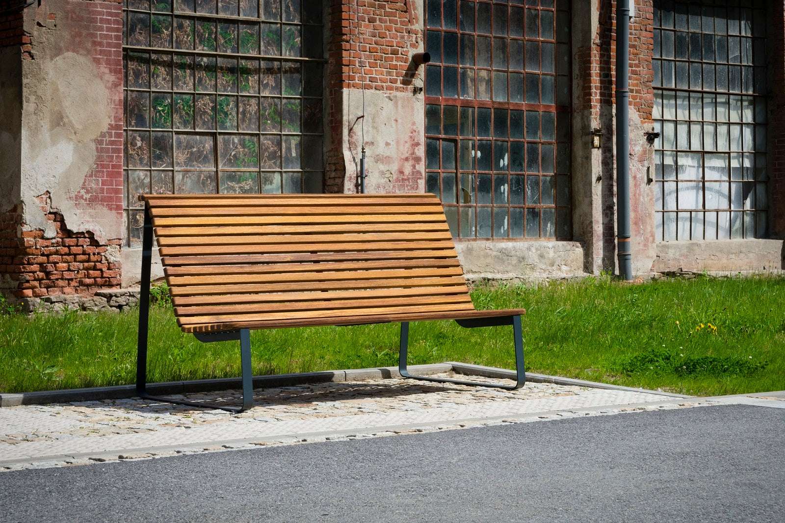 Moderne Holzparkbank mit Metallgestell steht auf einem gepflasterten Weg vor einer alten Backsteinfassade.