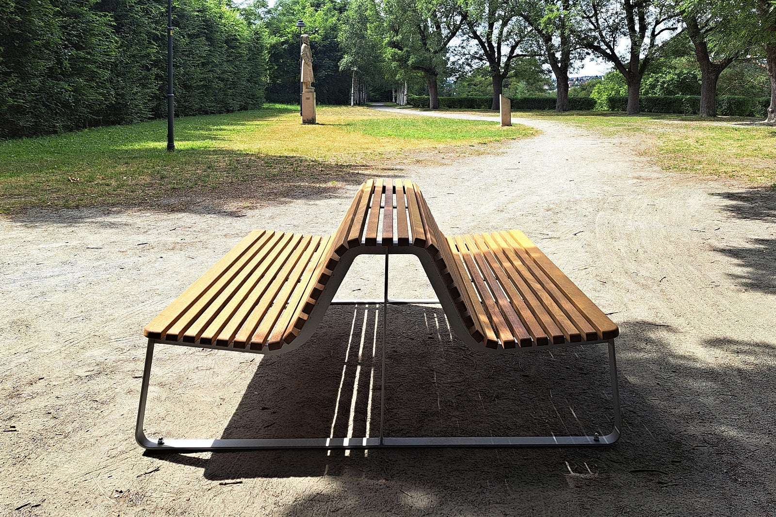 Moderne Holzparkbank mit Metallgestell auf Sandweg, umgeben von Bäumen im Park.