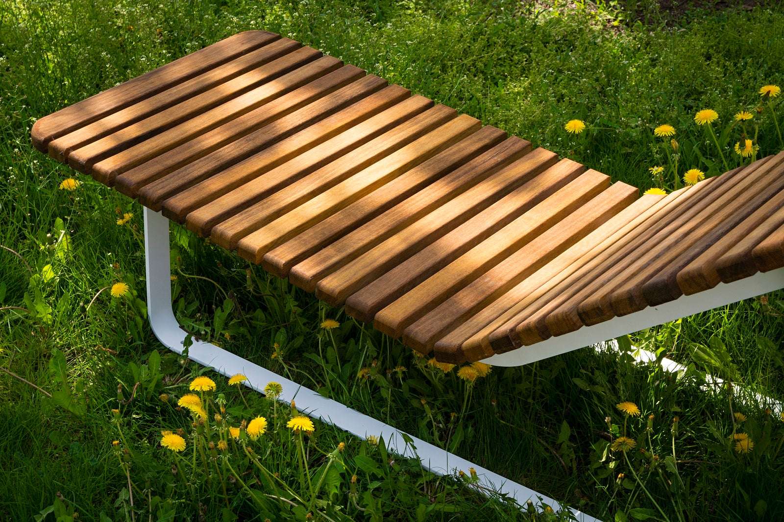 Moderne Holzgartenbank mit Metallgestell, umgeben von grüner Wiese und gelben Löwenzahnblüten, in der Sonne.