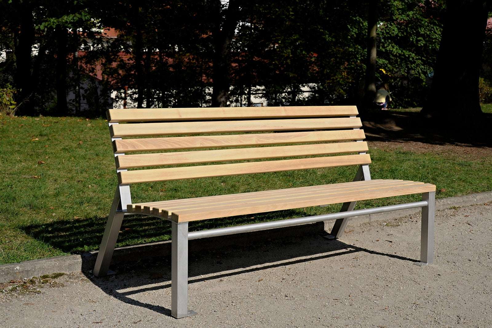 Hölzerne Parkbank mit Metallgestell auf Kiesweg, umgeben von grünem Rasen und Bäumen im Hintergrund.