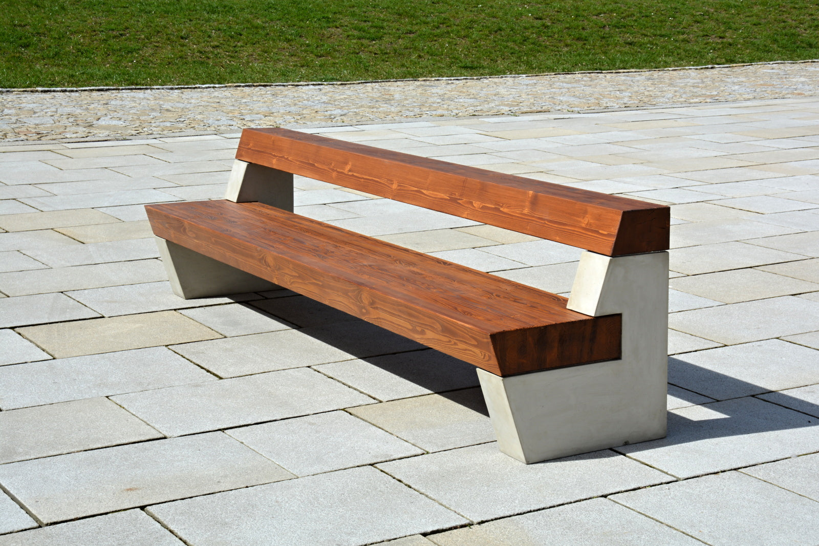 Moderne Bank mit Holzsitz und Lehne auf massiven Betonsockeln, auf einem gepflasterten Weg vor einer Grünfläche platziert.