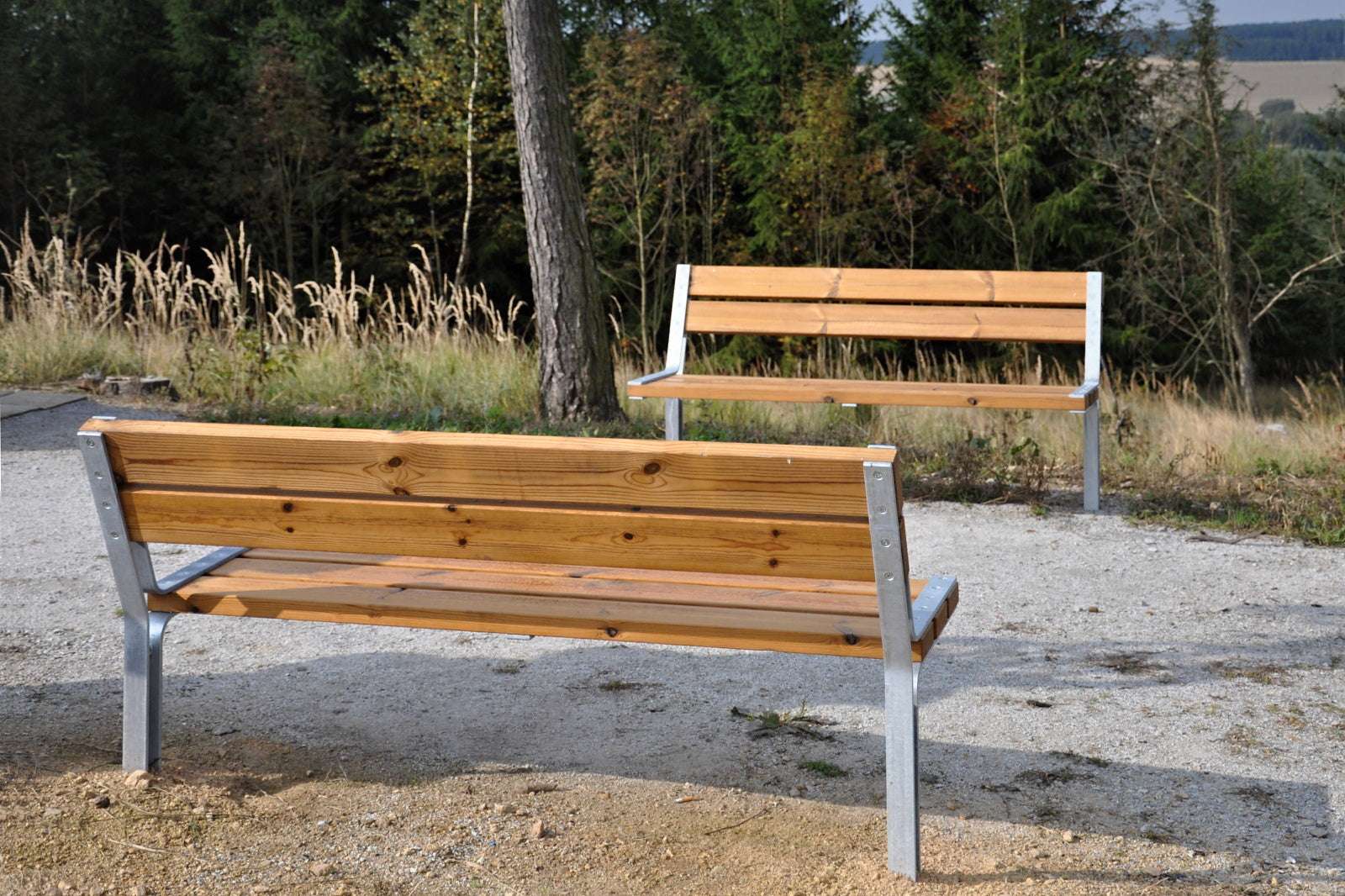 Zwei Holzbänke mit Metallgestell stehen auf einem Kiesweg in einer natürlichen, bewaldeten Umgebung bei Tageslicht.