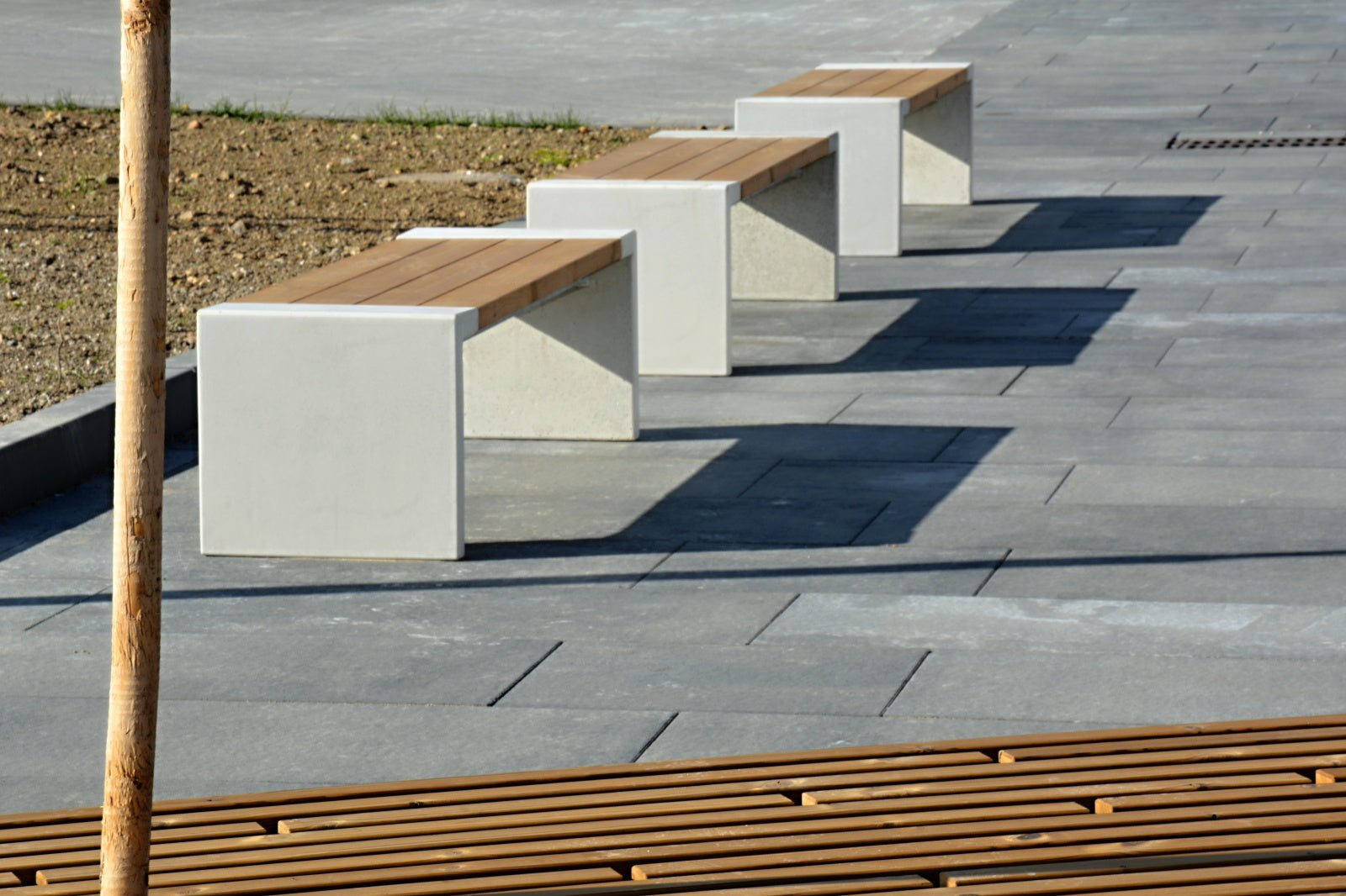 Auf dem Bild sind drei moderne Bänke mit weißen Betonsockeln und hölzernen Sitzflächen auf einem gepflasterten Platz zu sehen. Im Vordergrund ragt ein Baumstamm ins Bild, daneben befinden sich Holzlatten auf dem Boden. Im Hintergrund ist ein Bereich mit Erde erkennbar. Die Bänke werfen lange Schatten auf die grauen Bodenplatten. Die Umgebung wirkt urban und sauber, vermutlich eine öffentliche Fläche oder ein Park. Die Materialien der Bänke und des Bodens wirken hochwertig und gepflegt.