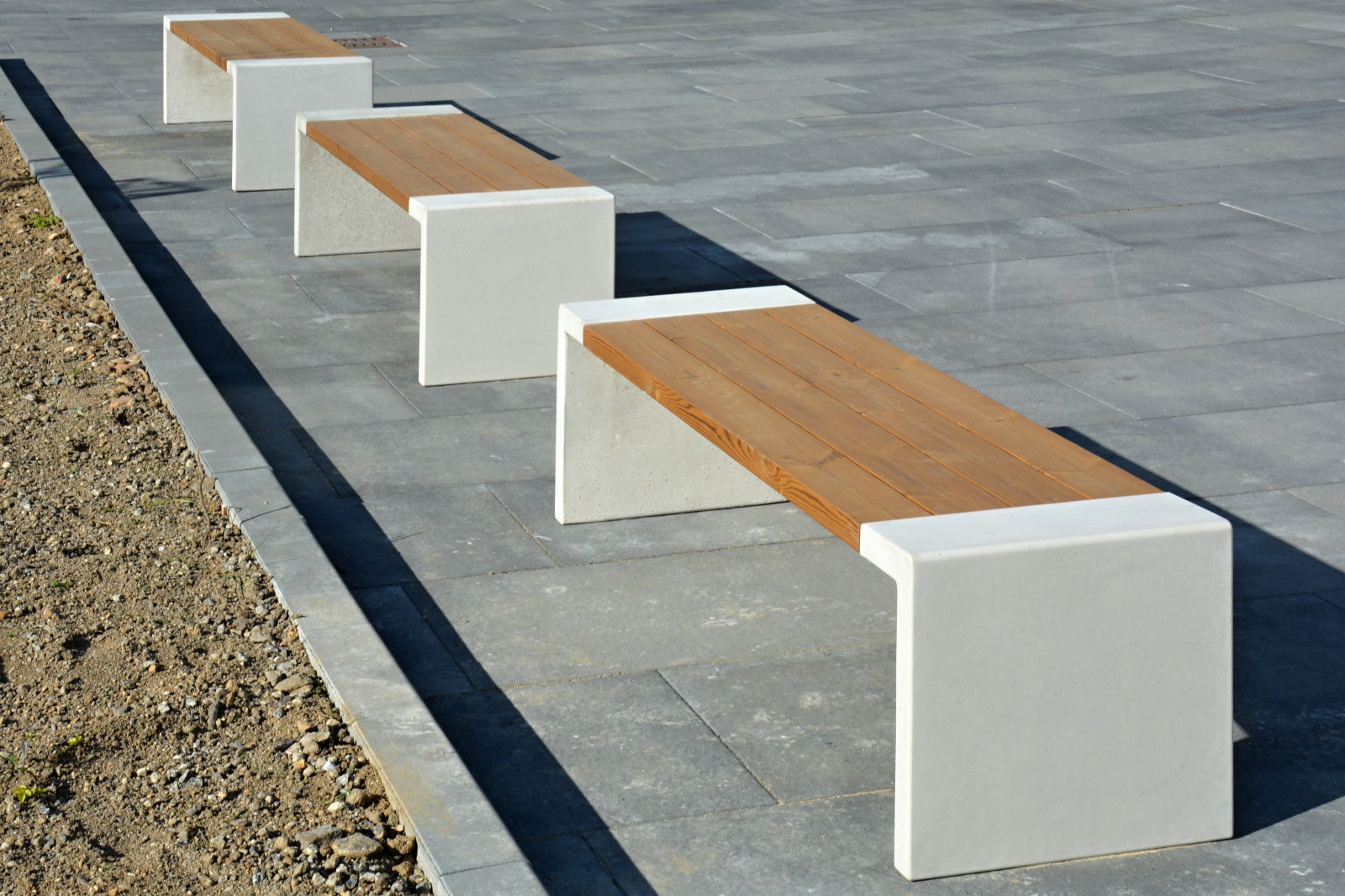 Drei moderne Bänke mit Sitzflächen aus Holzlatten und weißen, rechteckigen Betonstützen stehen auf einem gepflasterten Platz. Links befindet sich eine sandige Erdefläche, der Rest ist von großen, grauen Steinplatten bedeckt. Die Bänke stehen in einer geraden Reihe und werfen deutliche Schatten auf den Boden. Es sind keine weiteren Gegenstände oder Personen sichtbar, die Umgebung wirkt offen und urban.
