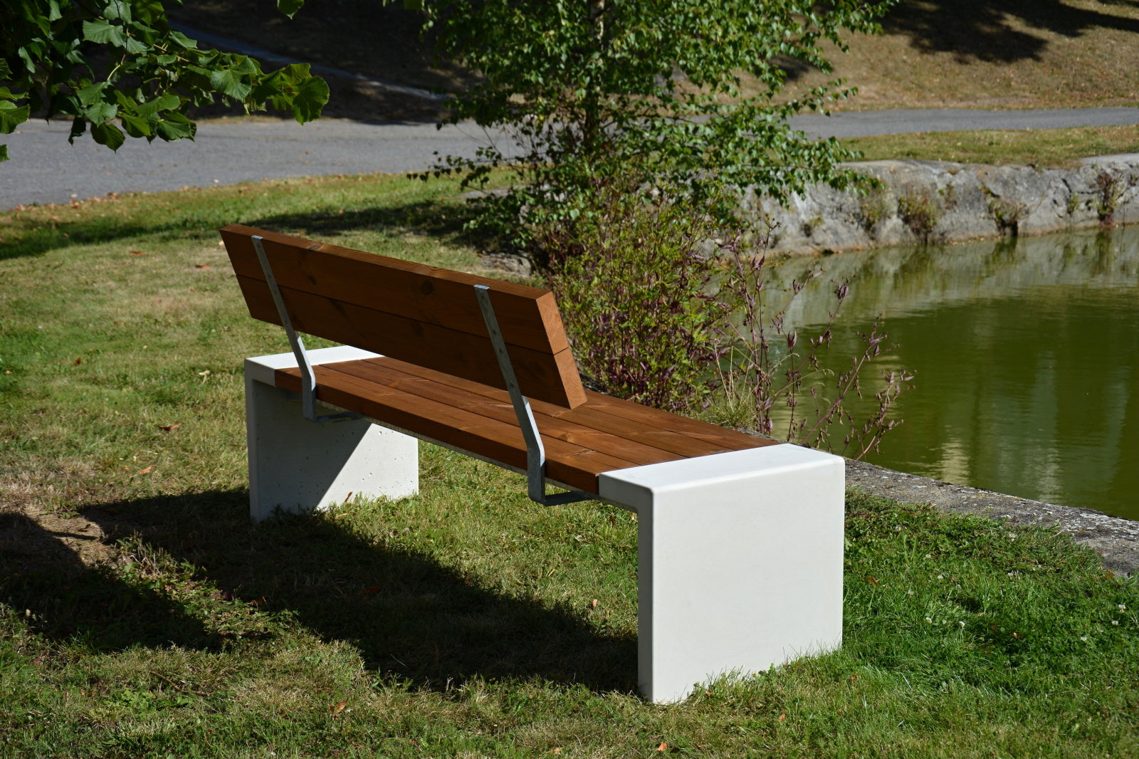 Holz-Sitzbank mit Betonfüßen am Teichufer in grüner Parklandschaft.