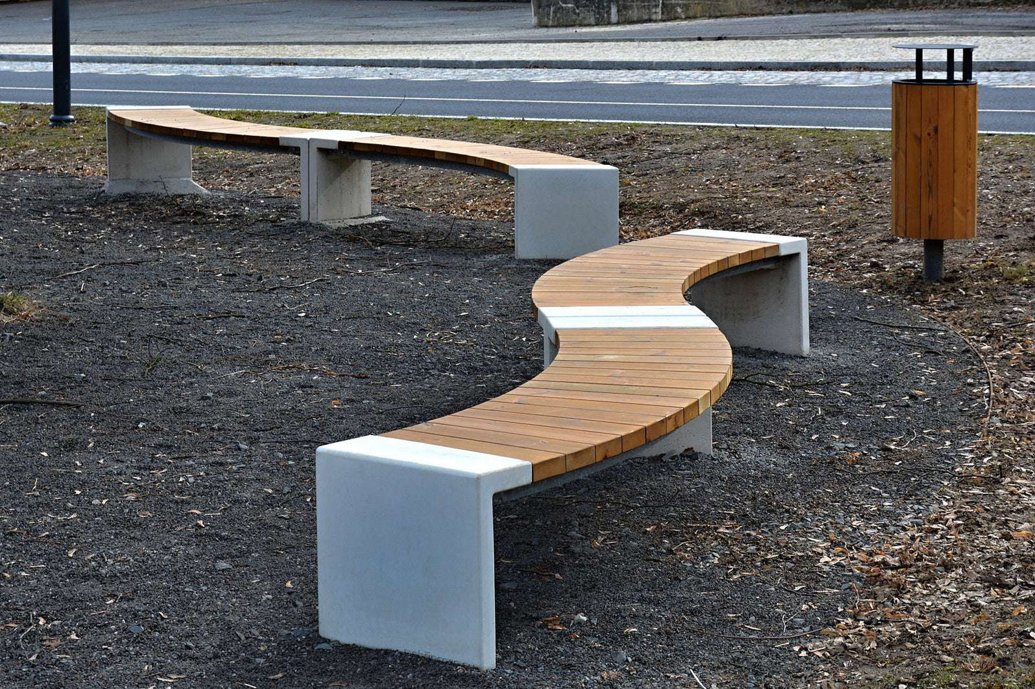 Geschwungene Parkbank aus Holz und Beton auf grauem Kies mit Holz-Mülleimer im Hintergrund.