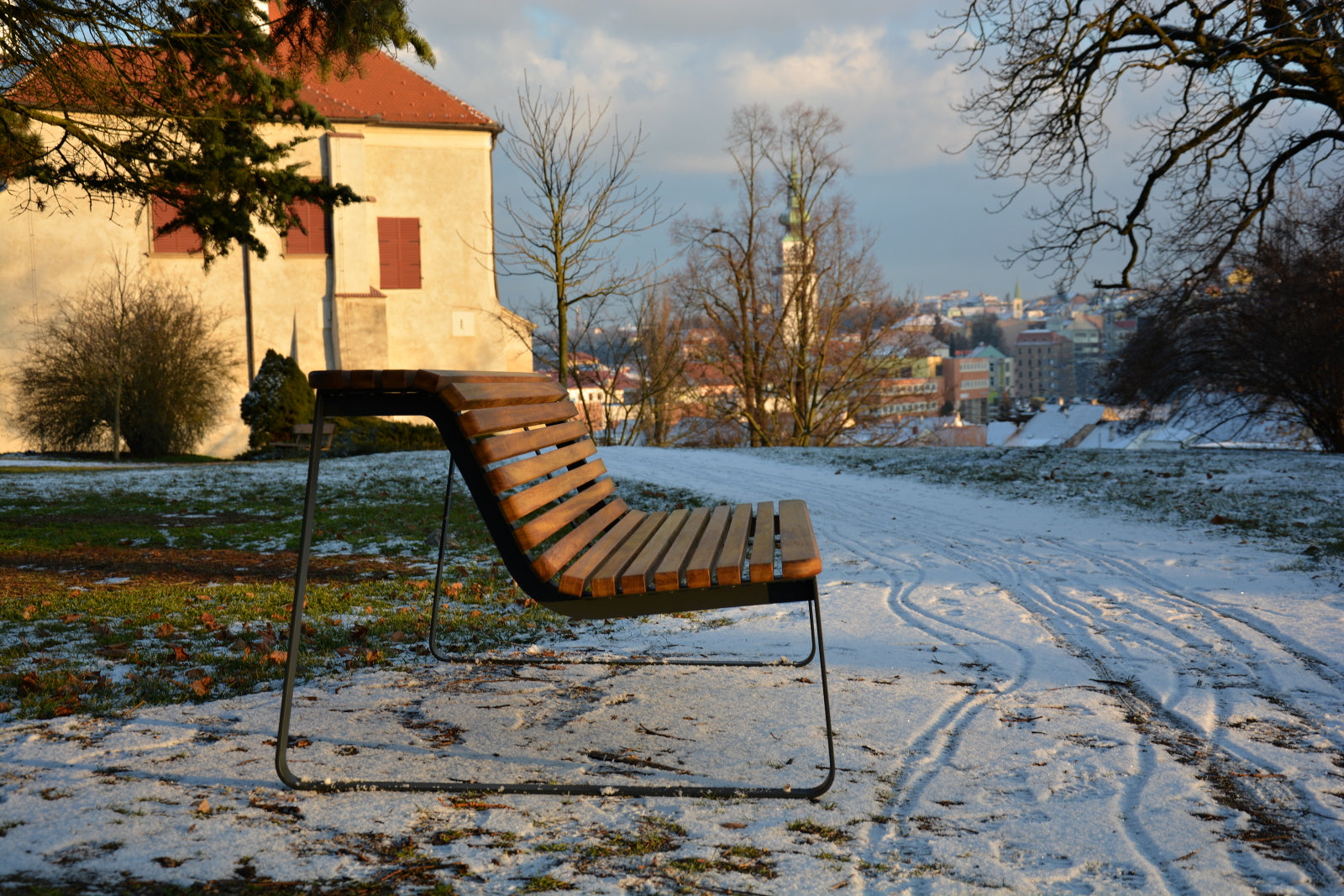 Moderne Holzparkbank auf verschneitem Weg, mit Aussicht auf historische Gebäude im Hintergrund.