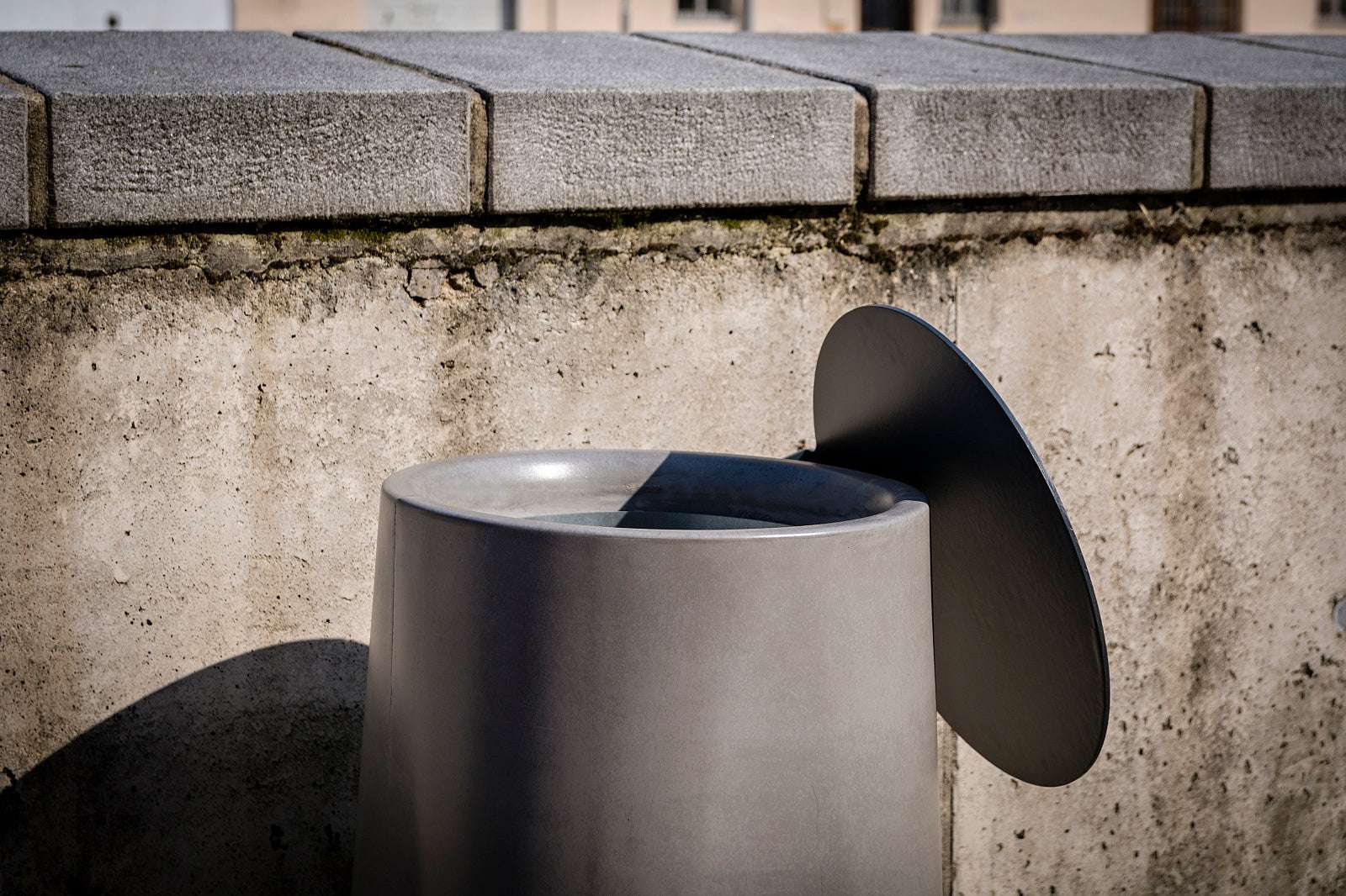 Moderner, grauer Abfallbehälter aus Metall vor einer Betonwand, teils im Schatten.
