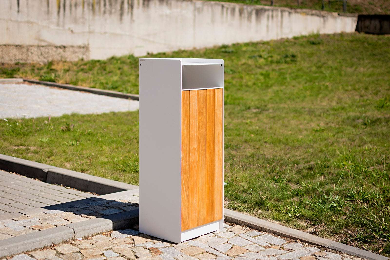 Mülleimer mit weißem Metallrahmen und hölzerner Front steht auf gepflastertem Weg im Freien.