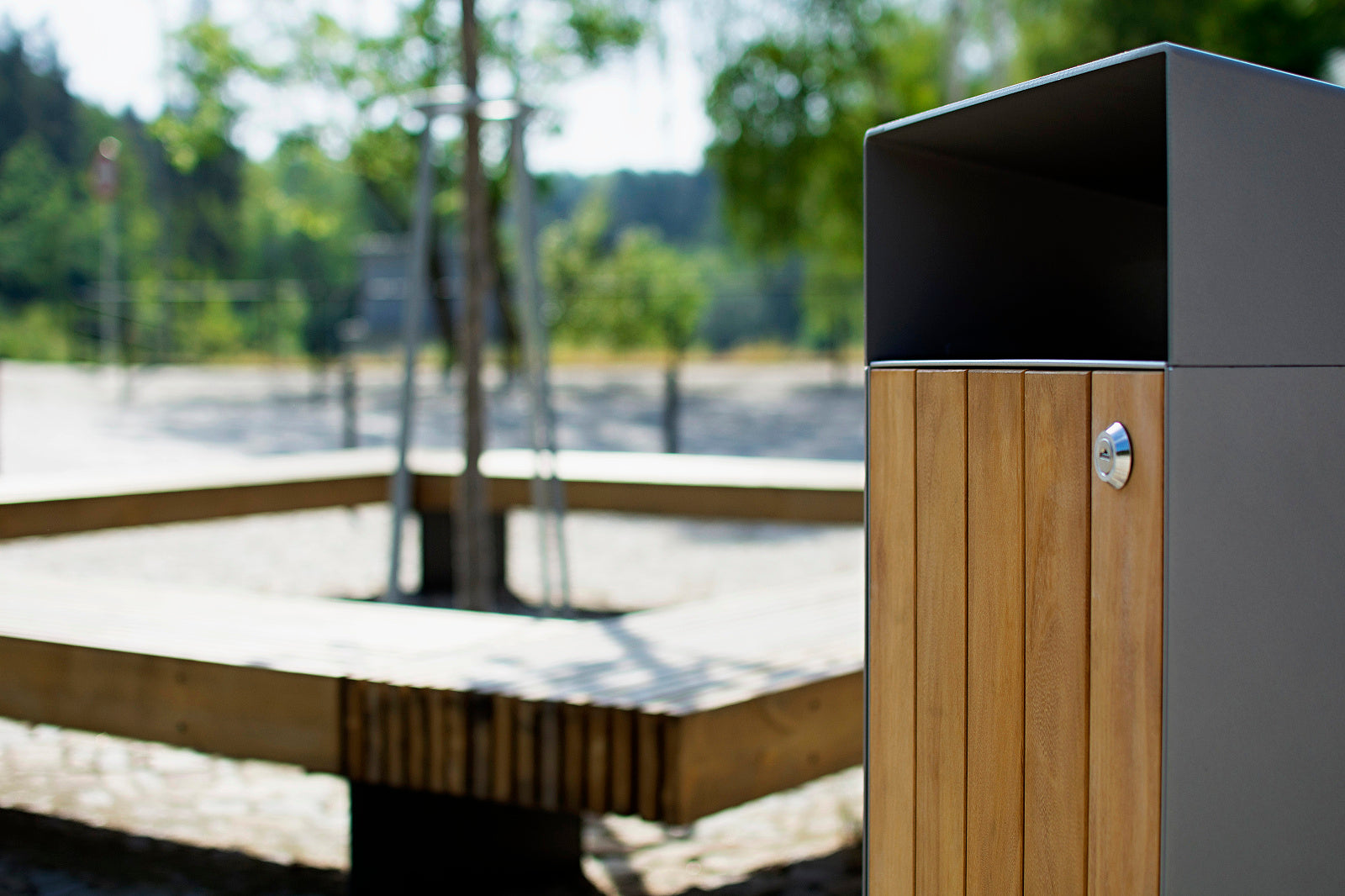 Im Vordergrund steht ein moderner Abfallbehälter mit vertikalen Holzleisten und einem Metall-Gehäuse sowie Schloss. Im Hintergrund befindet sich eine eckige Sitzbank aus Holz, einige junge Bäume mit Stützpfählen und ein weitläufiger, sonniger Platz mit sandigem Untergrund. Weiter hinten sind undeutliche Bäume und Vegetation sichtbar. Die Szene wirkt wie ein Park oder öffentlicher Außenbereich.
