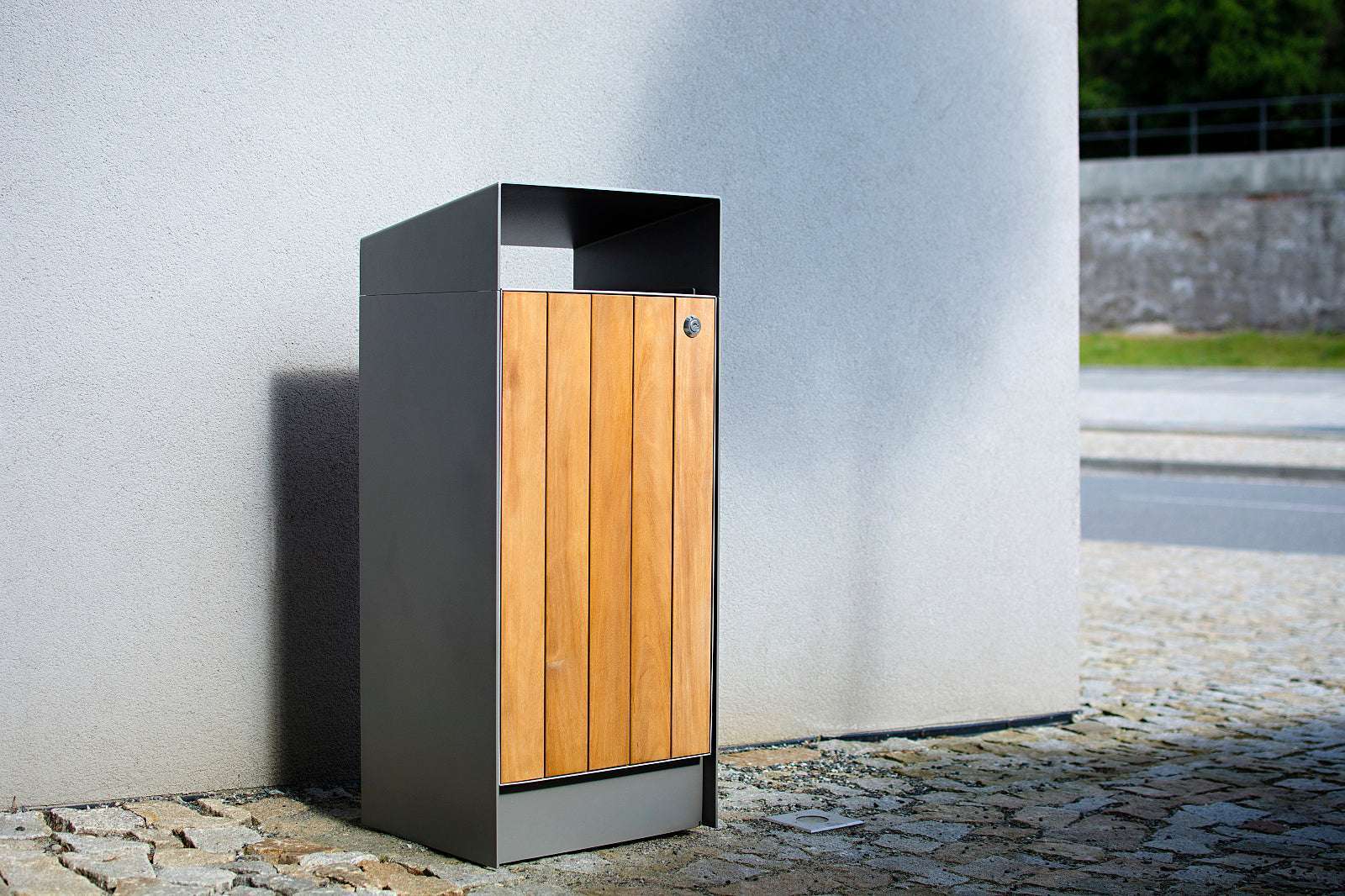 Moderner Abfallbehälter mit Holzverkleidung auf gepflasterter Fläche, neben grauer Wand platziert.