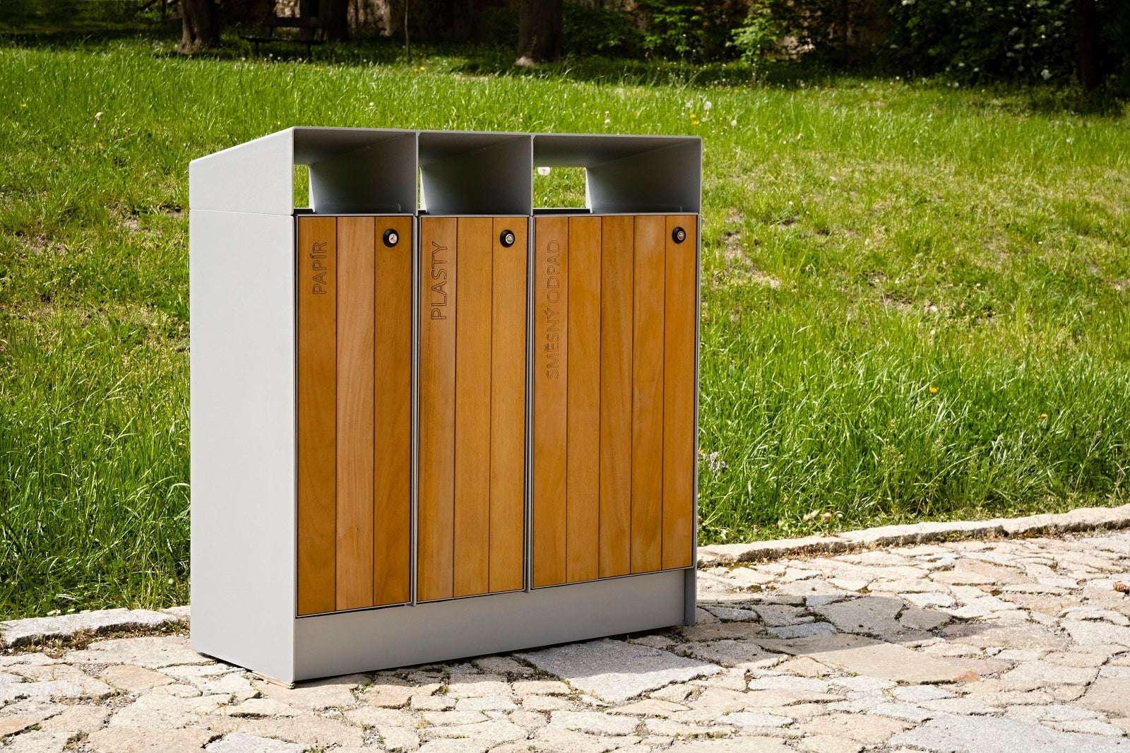 Moderner Abfallbehälter aus Holz und Metall mit drei Fächern für Papier, Plastik und Glas im Freien.