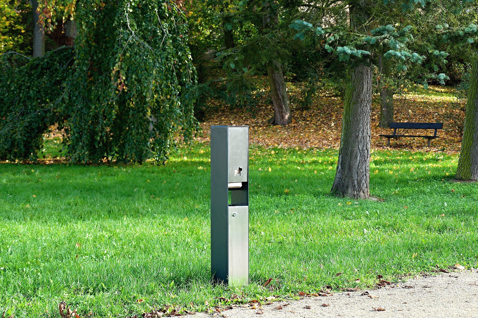 Ein moderner, grauer Mülleimer steht in einem grünen Park mit Bäumen und Laub im Hintergrund.