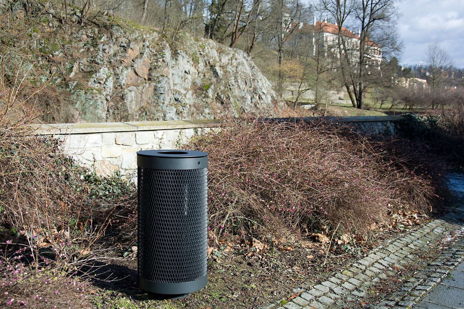 Ein schwarzer Metall-Abfallbehälter steht auf einem gepflasterten Weg, umgeben von kahlen Büschen und Natur.