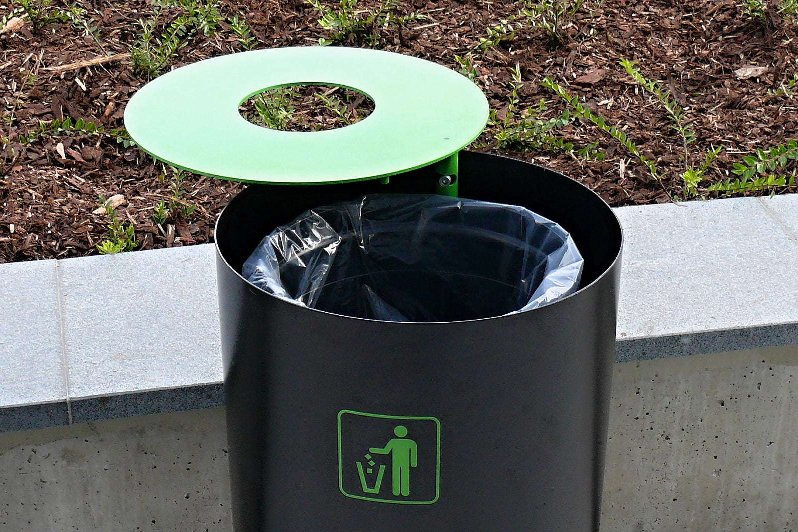 Schwarzer Mülleimer mit grünem Deckel und Symbol, aufgestellt vor einer Mauer mit Pflanzen im Hintergrund.