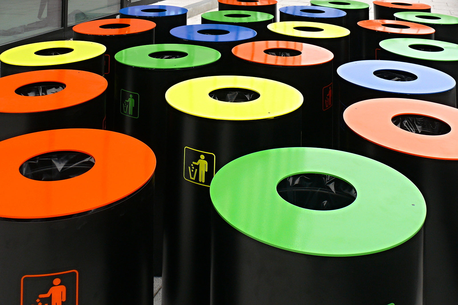 Mehrere schwarze Abfallbehälter mit großen, runden Öffnungen sind aufgereiht. Die Deckel sind in kräftigen Farben gestaltet: Orange, Gelb, Grün und Blau. Auf der Vorderseite der Behälter befindet sich ein Symbol eines Menschen, der Müll wegwirft, oft passend zur Deckelfarbe umrandet. Im Hintergrund spiegeln Fenster die Szene wider. Im Inneren der Behälter sieht man schwarze Müllbeutel. Die Behälter stehen dicht beieinander auf einem Boden aus grauen Fliesen.