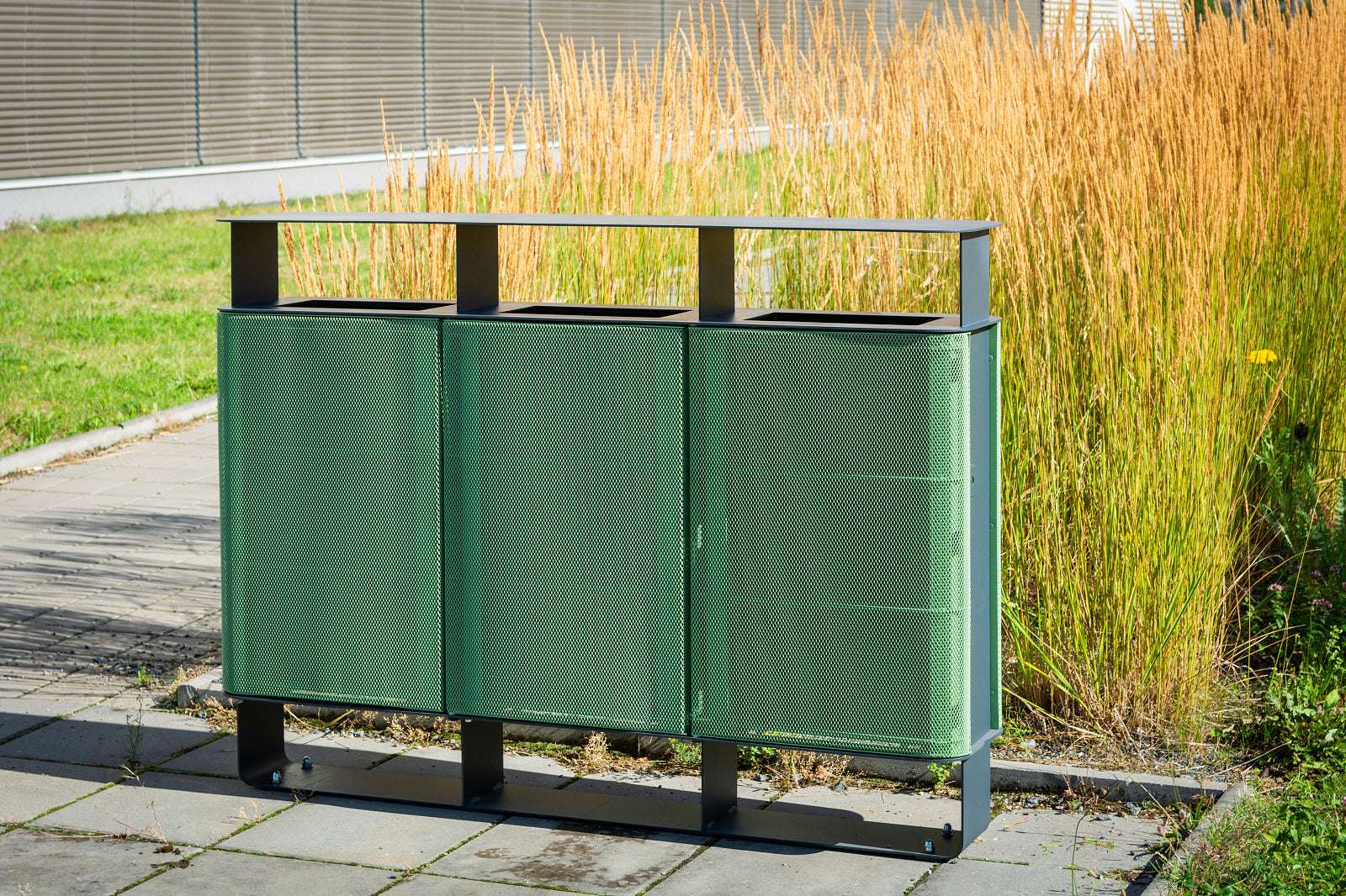 Moderner grüner Stadion-Abfallbehälter mit drei Fächern, aufgestellt neben hohem Gras auf gepflastertem Boden.