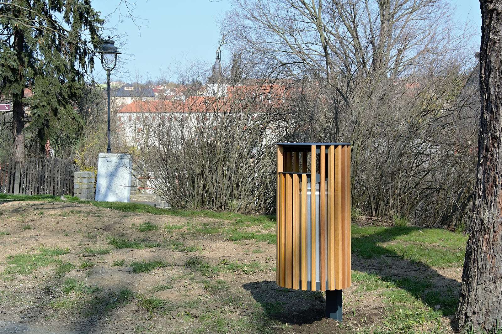 Holzvertäfelter Abfallbehälter im Park, umgeben von Bäumen und Büschen, mit Häusern im Hintergrund.