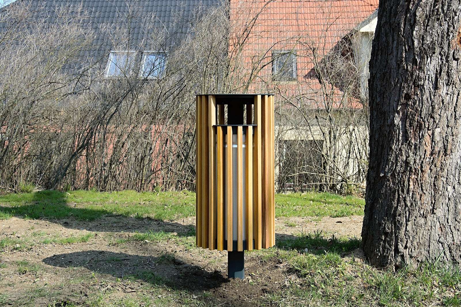 Moderner Abfallbehälter aus Holz und Metall, steht im Freien neben einem Baum, mit Wohngebäude im Hintergrund.