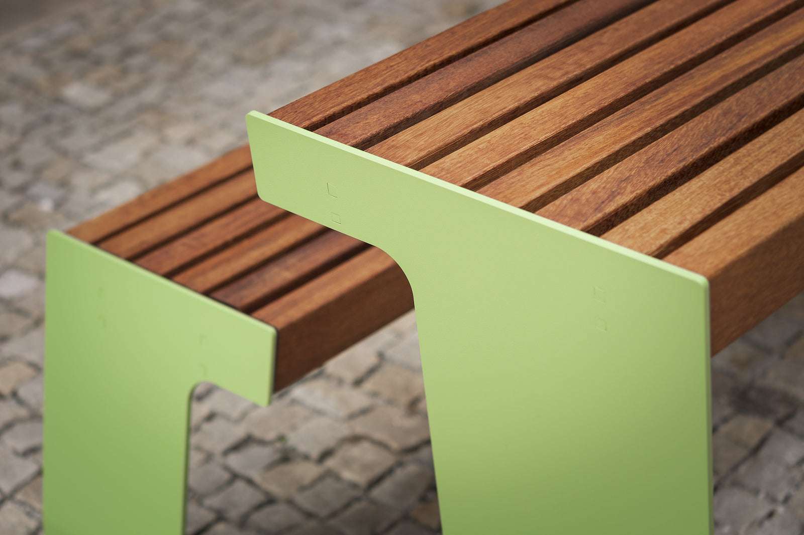 Moderne Holzbank mit grünem Metallrahmen auf einem Kopfsteinpflaster.