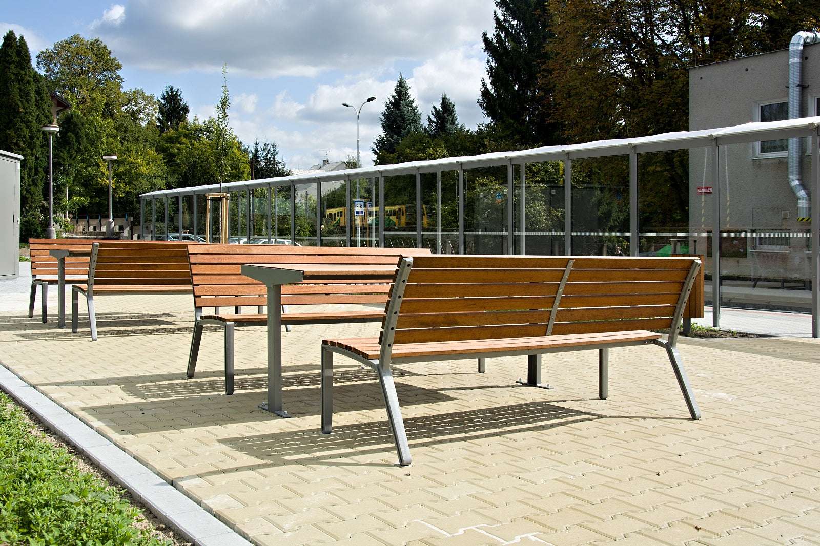 Holzbänke auf gepflastertem Platz mit Glasschutzwand im Freien, umgeben von Bäumen und Gebäuden.