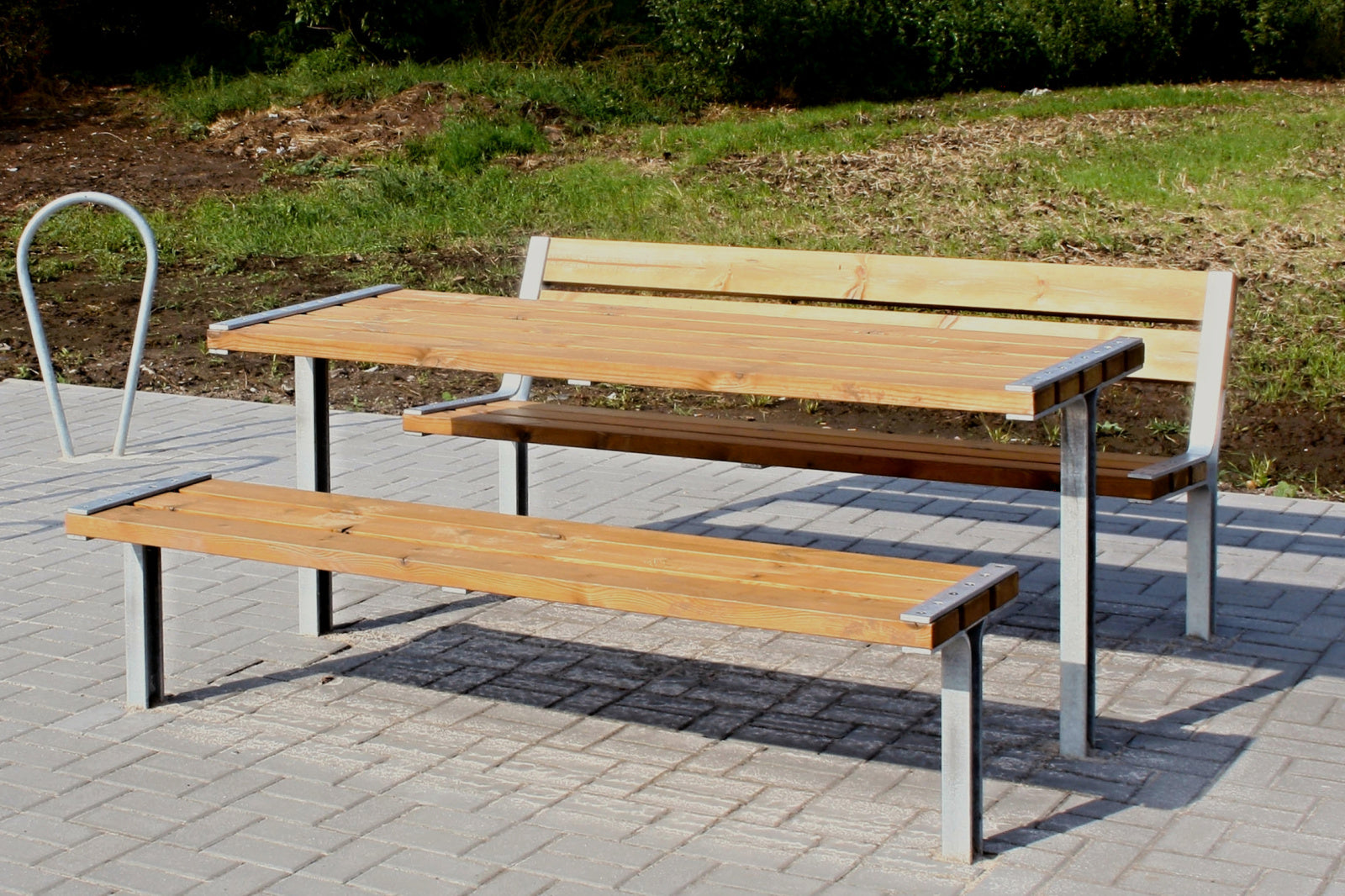 Holz-Sitzgruppe im Freien mit zwei Bänken und Tisch auf gepflastertem Boden, ideal für Picknicks oder Pausen im Grünen.