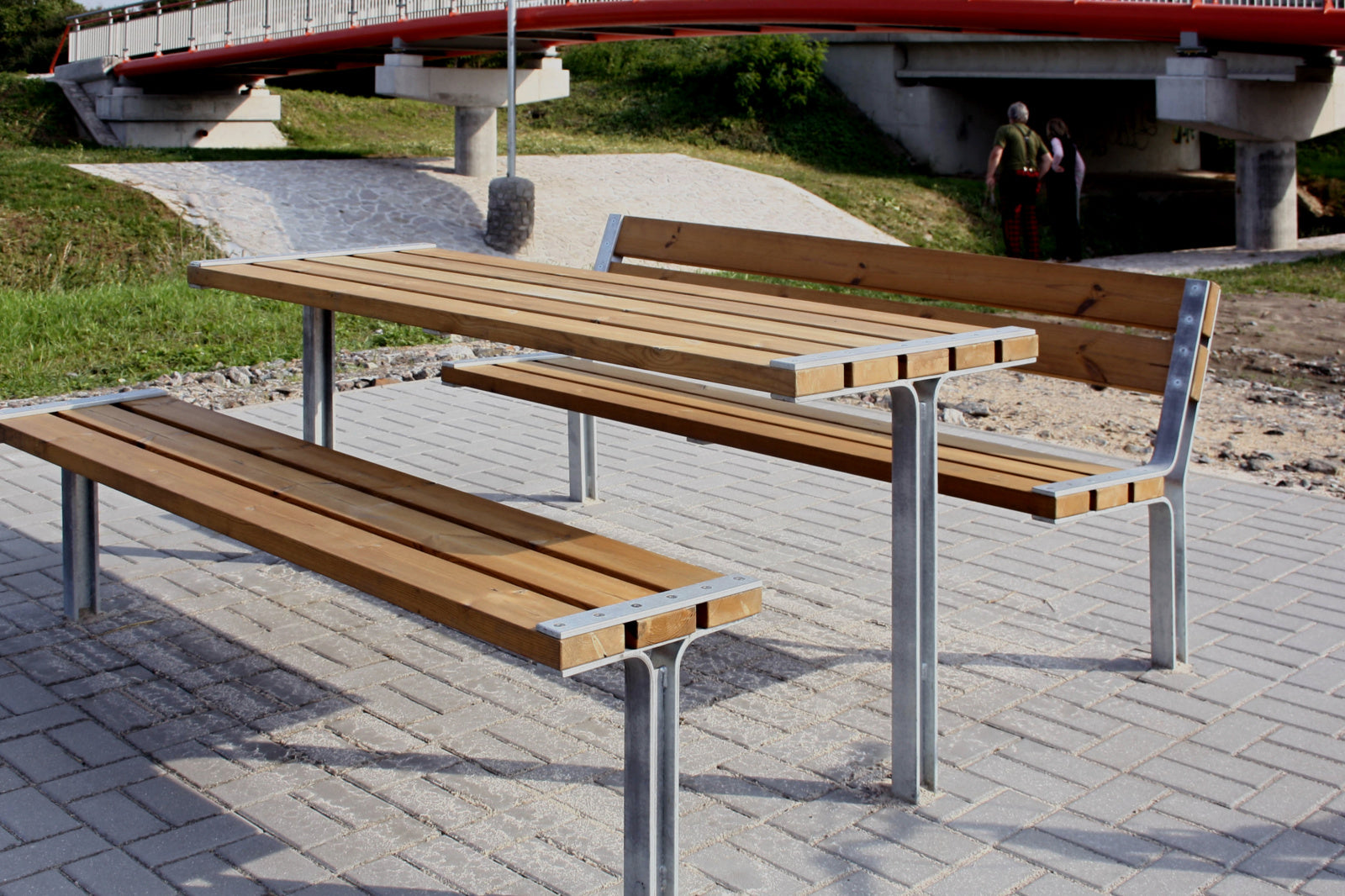 Eine moderne Outdoor-Sitzgruppe aus Metall und Holz, bestehend aus einem Tisch und zwei Bänken, steht auf Pflastersteinen.
