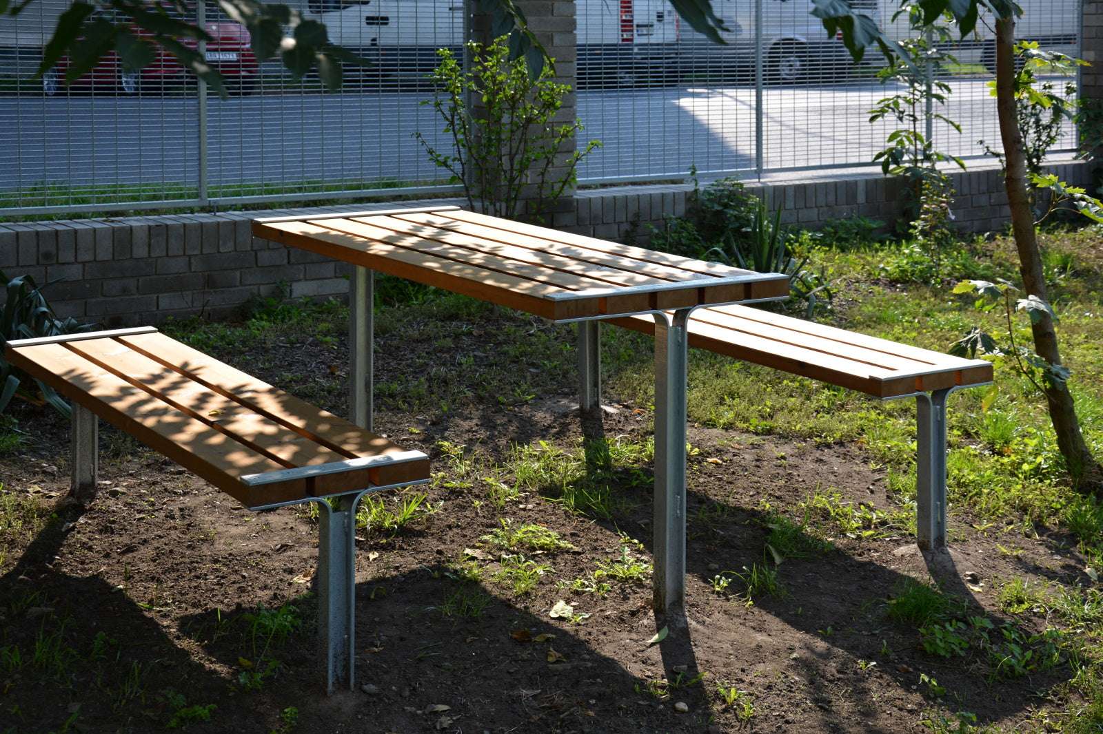 Garten-Tisch-und-Bank-Set aus Holz und Metall auf Grasfläche vor einem Zaun.