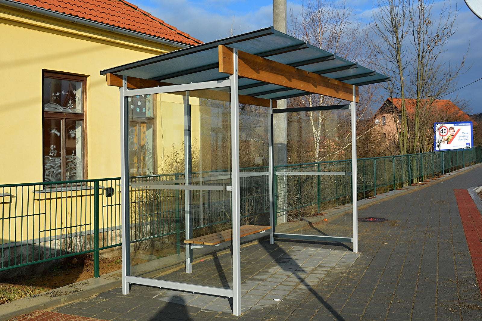 Bushaltestelle mit Glaswänden und Holzdach neben gelbem Gebäude.