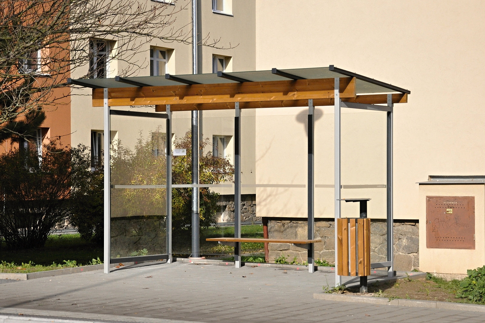 Eine moderne Bushaltestelle mit Glaswänden, einem Holzdach und einer Holzsitzbank, umgeben von Häusern und Grünflächen.