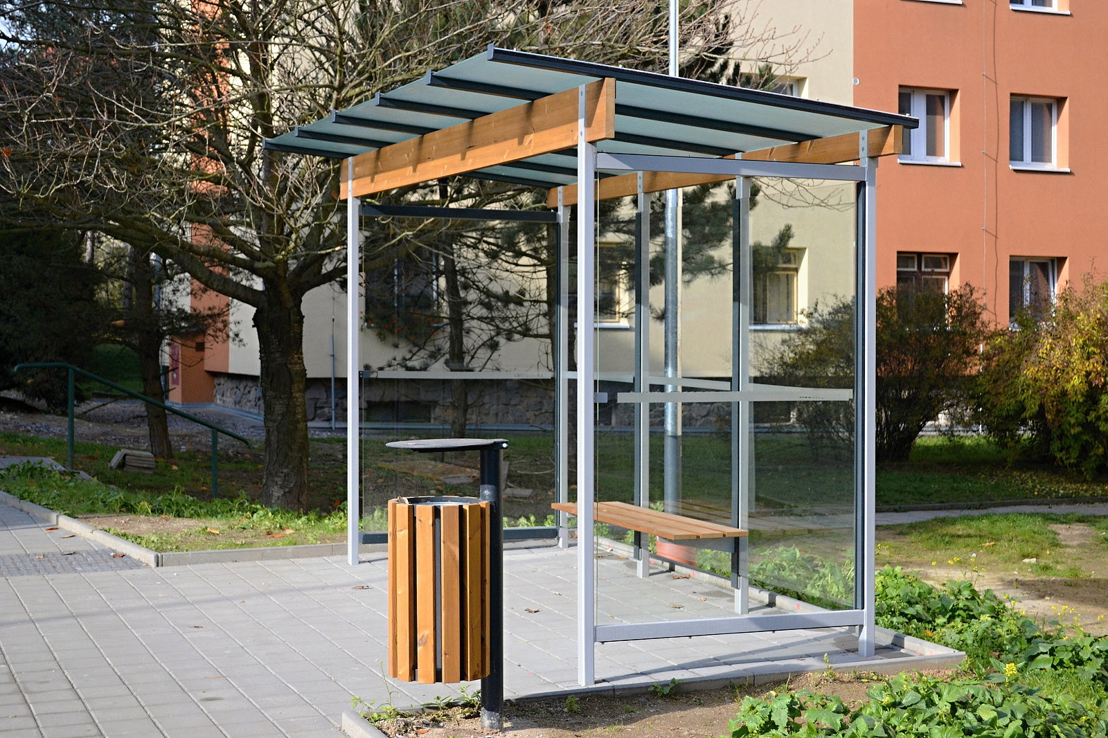 Auf dem Bild ist eine moderne Wartehalle mit Glasdach und Seitenwänden, integriertem Holzsitz und Abfalleimer auf einem Gehweg.