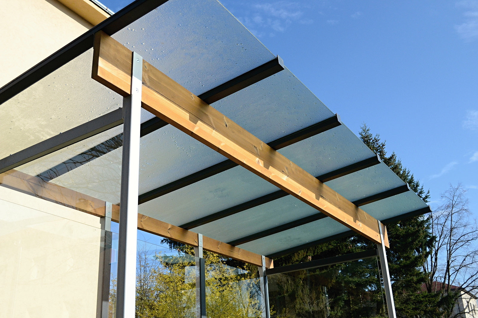 Das Bild zeigt ein modernes Vordach oder eine Überdachung aus Glasplatten und Holzbalken, gestützt von Metallpfosten. Im Hintergrund sind Bäume und blauer Himmel zu sehen. Die Glasflächen weisen Wassertropfen auf, was auf Feuchtigkeit oder Regen hindeutet. Die Konstruktion wirkt stabil und ist an der Seite eines Gebäudes angebracht. Das Glasdach lässt Licht durch und bietet gleichzeitig Schutz.