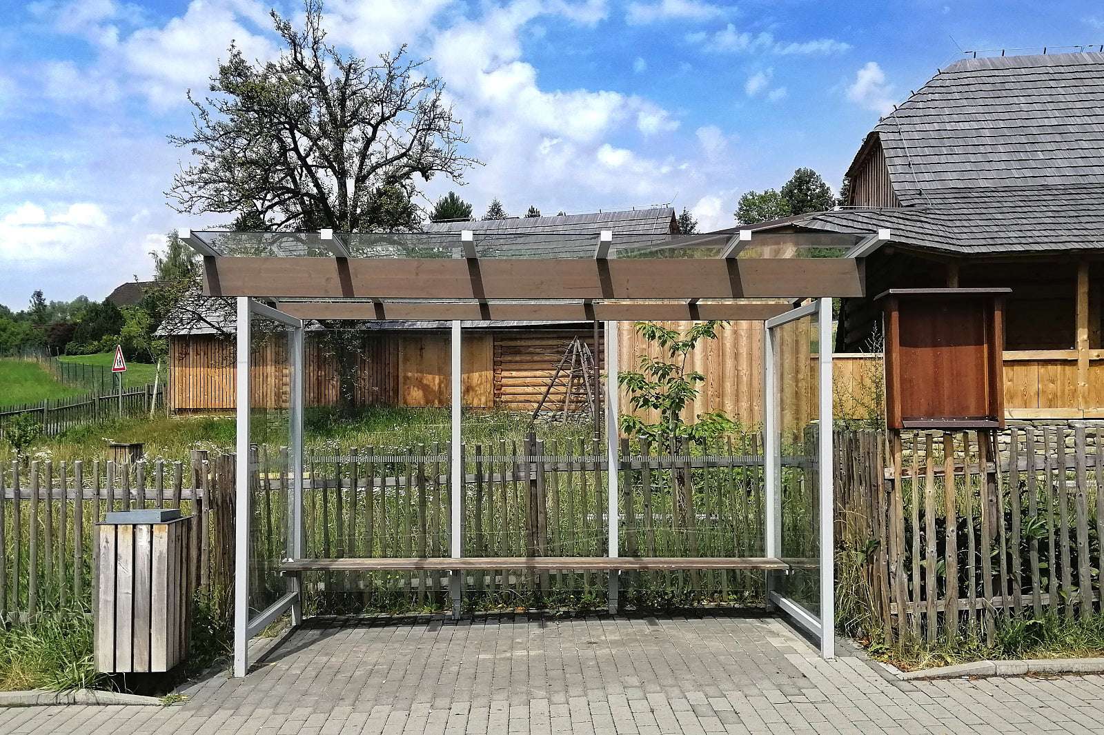 Bushaltestelle mit Überdachung aus Glas und Metall, Holzhütten im Hintergrund, baumreiche Landschaft.