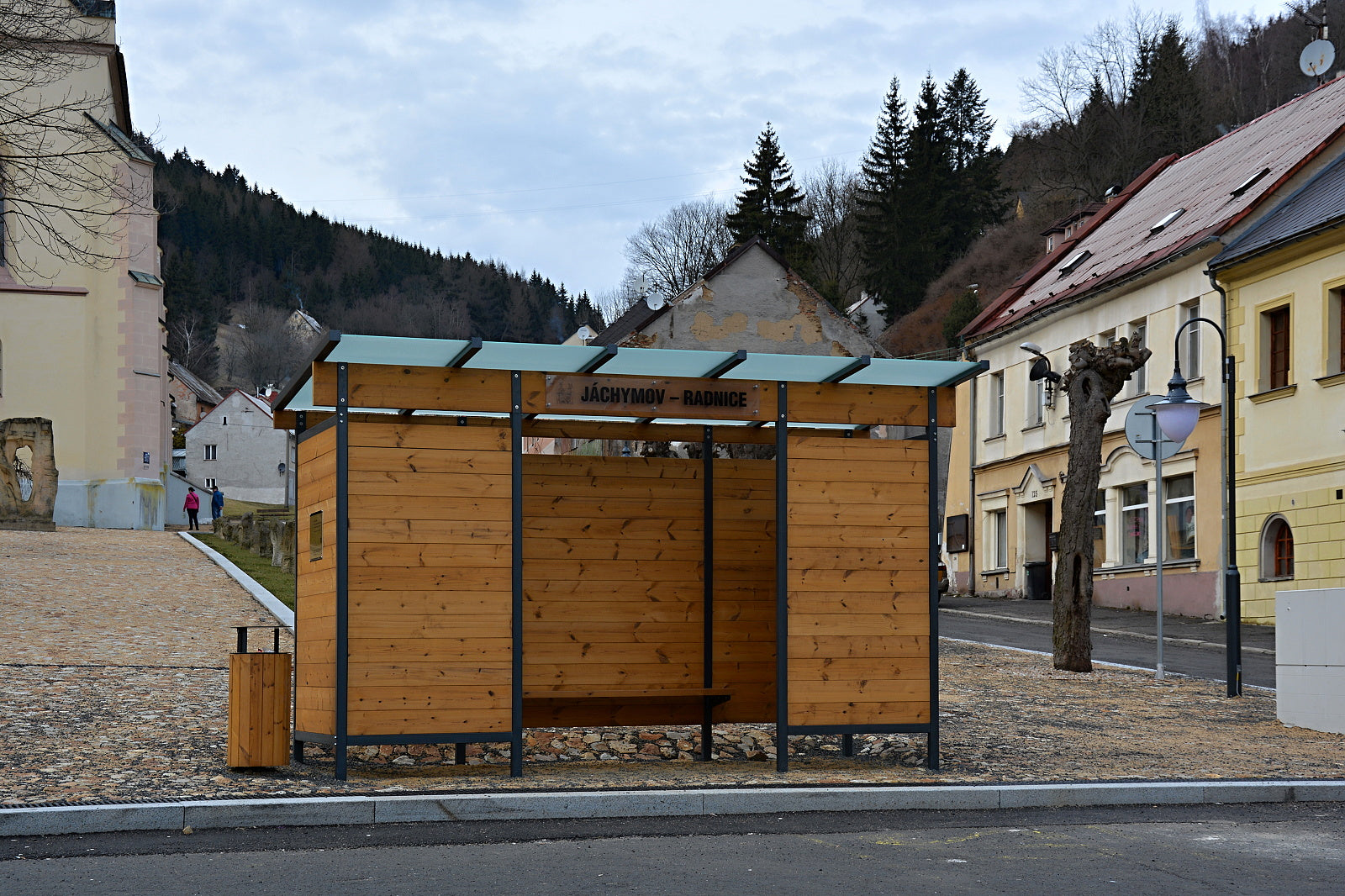 Holz- und Metallwartehalle mit Sitzbank und Mülleimer auf gepflastertem Platz, umgeben von Gebäuden und bewaldeten Hügeln.
