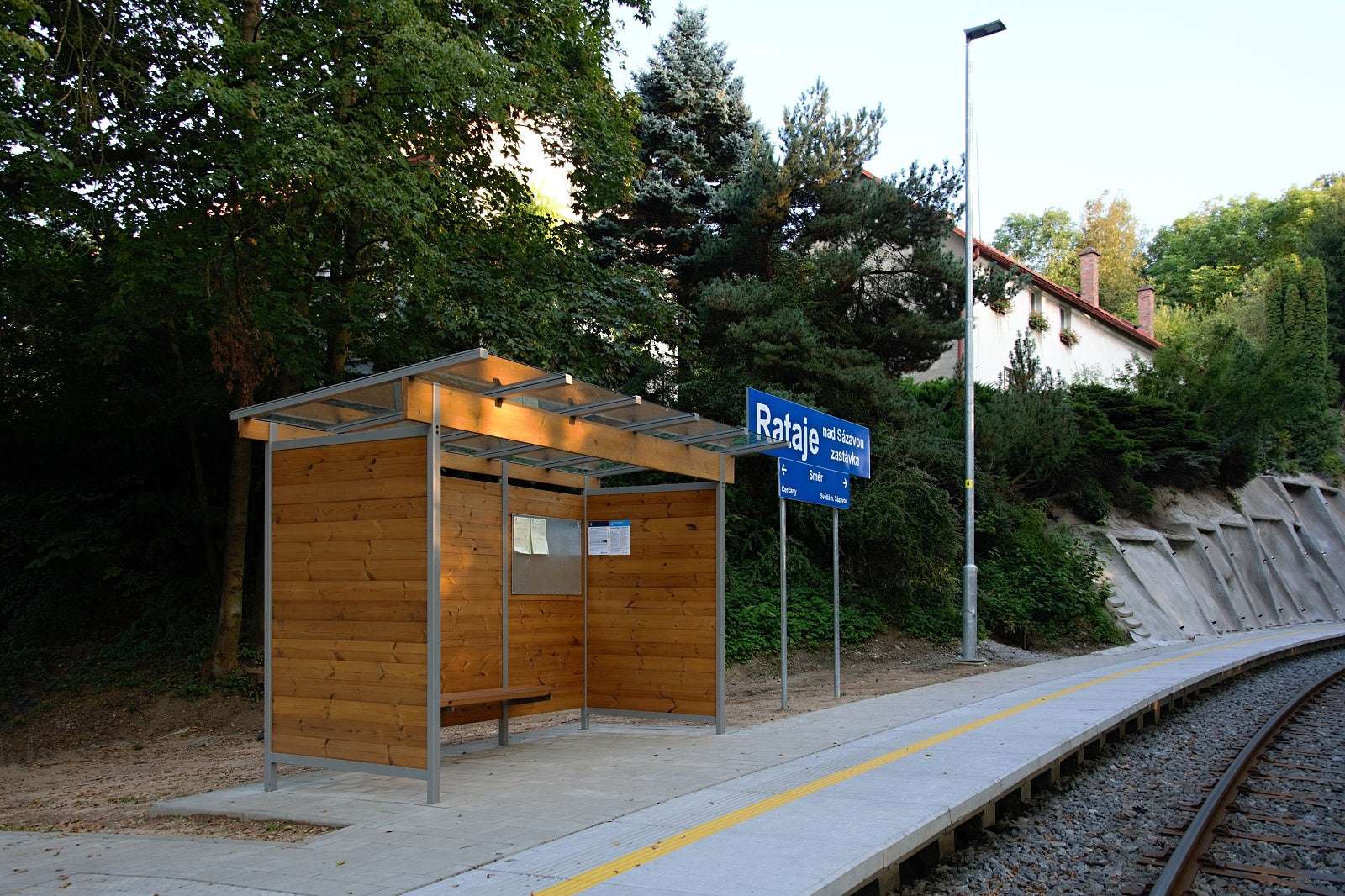 Holz-Wartehalle an Bahngleis mit Glasdach, Sitzbank und Beleuchtung, umgeben von Bäumen und Beschilderung.