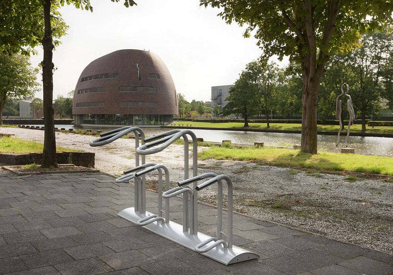Im Vordergrund steht ein moderner silberner Fahrradständer auf grauen Pflastersteinen. Im Hintergrund ist ein kleiner Fluss oder Teich zu sehen, daneben grüner Rasen und mehrere Bäume. Links sieht man ein auffälliges, rundes, kuppelartiges Gebäude mit Backsteinfassade und schmalen Fensterschlitzen. Im rechten Hintergrund steht eine abstrakte menschliche Skulptur, vermutlich aus Metall. Die Szene wirkt ruhig und ist vermutlich Teil eines Campus oder Parks.