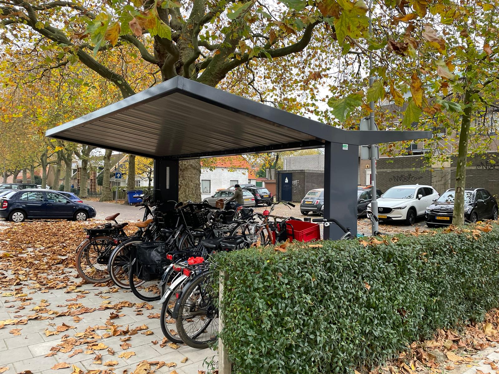 Überdachter Fahrradunterstand mit vielen abgestellten Fahrrädern, daneben Autos auf Parkplatz, Bäume mit Herbstlaub, niedrige Hecke vorne