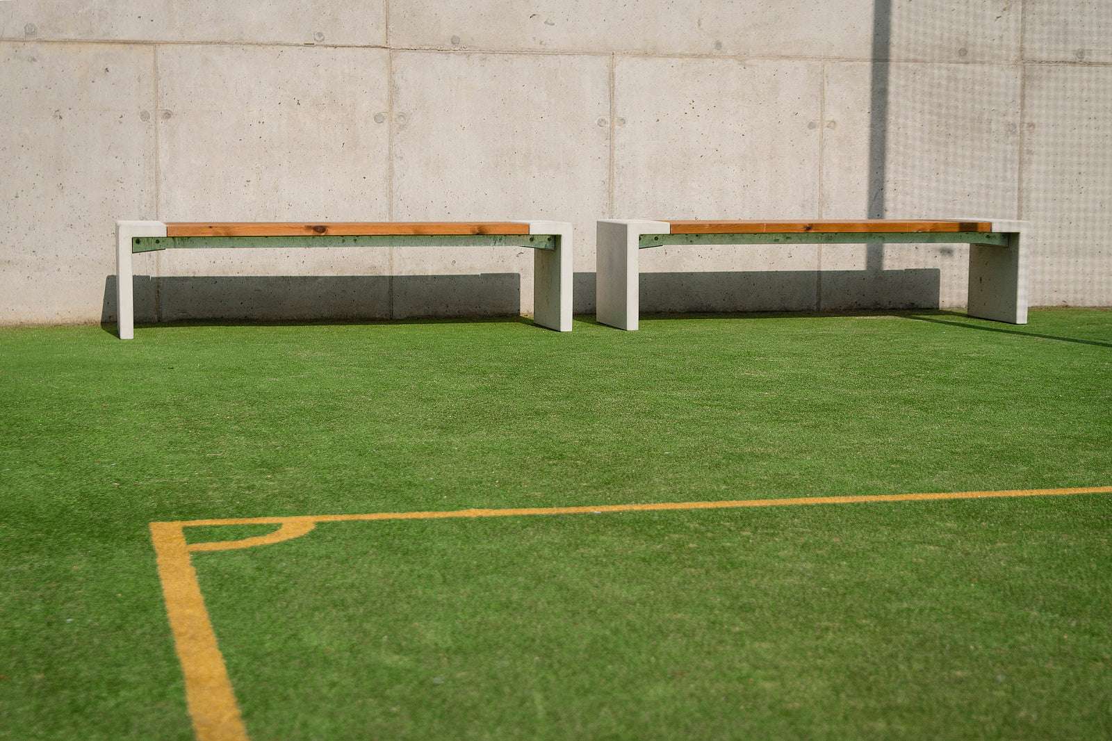 Zwei moderne Bänke auf Kunstrasen vor einer Betonwand, ideal für Außenbereiche und Sportplätze.