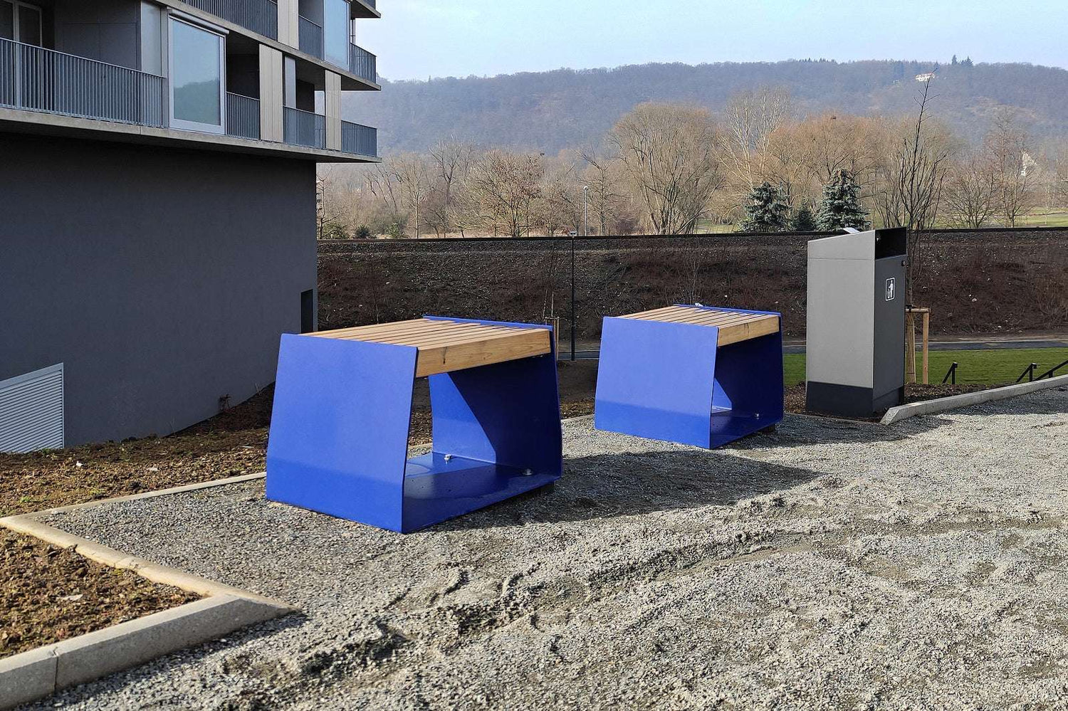 Blaue, moderne Outdoor-Bänke auf einem Kiesplatz neben einem Gebäude, mit Bäumen im Hintergrund.