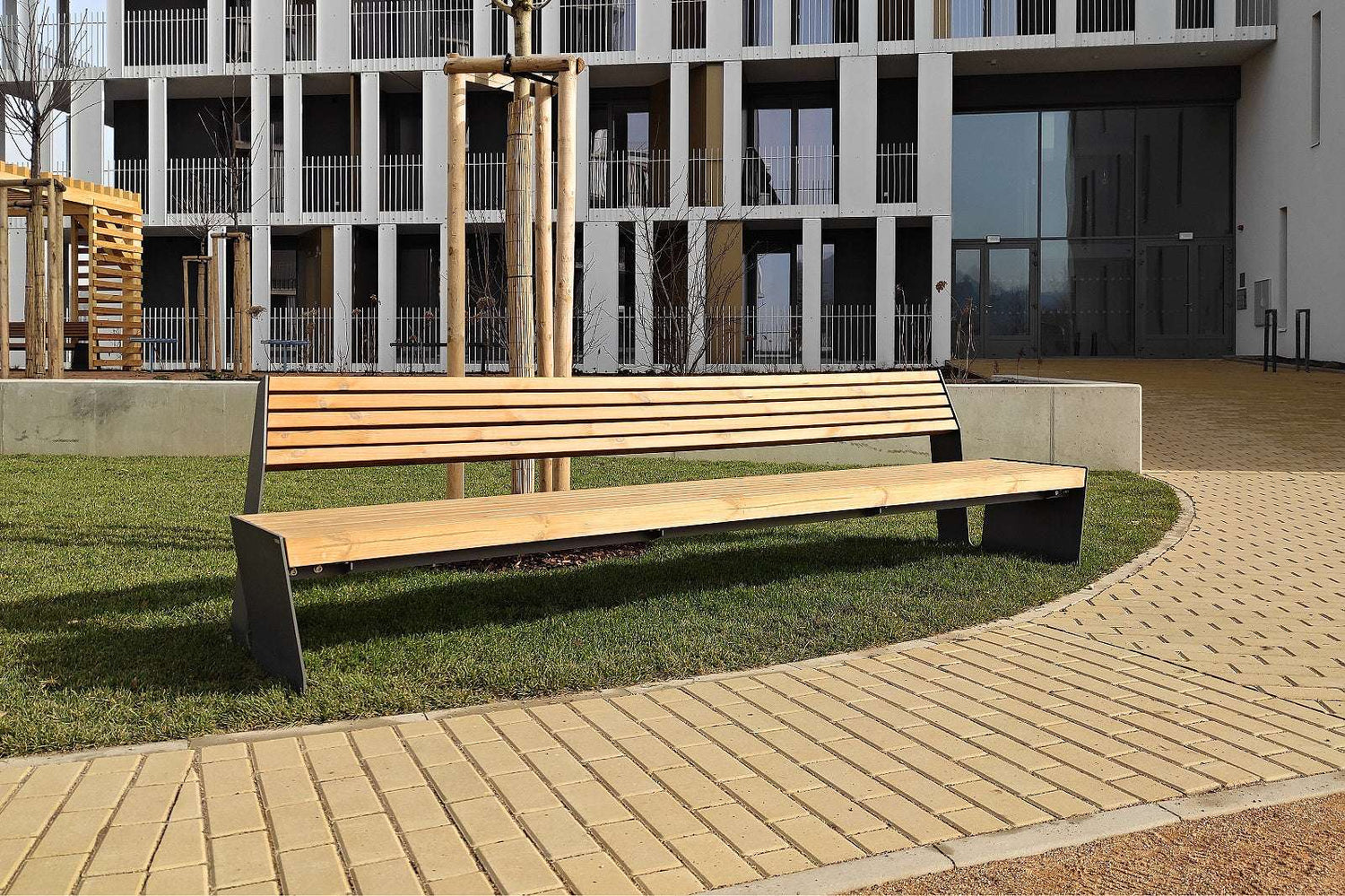 Moderne Holzbank mit Metallgestell auf gepflastertem Weg vor einem Gebäude mit Glasfassade.