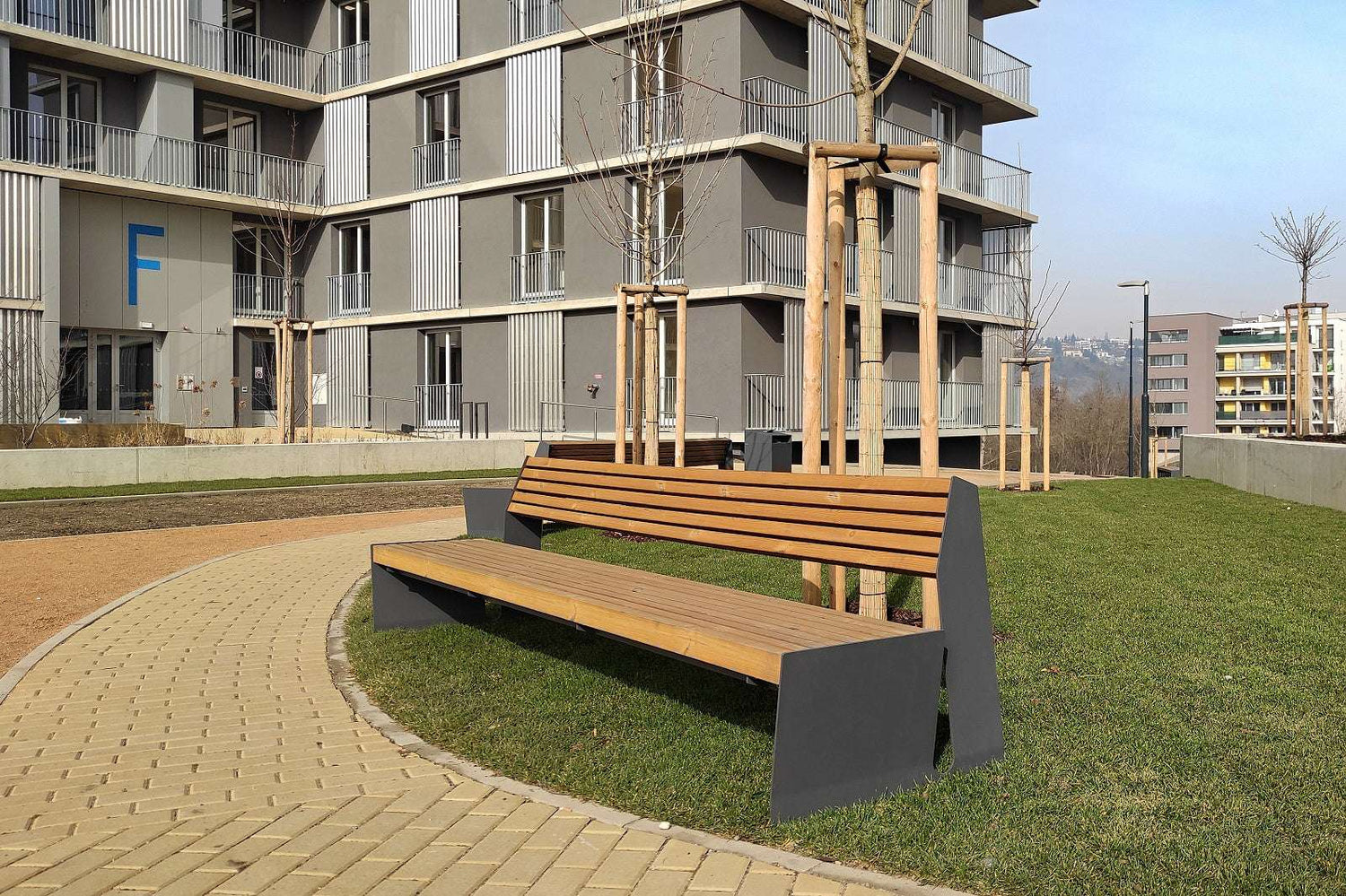 Moderne Holzbank auf einem gepflasterten Weg in einem städtischen Park vor einem Wohngebäude.