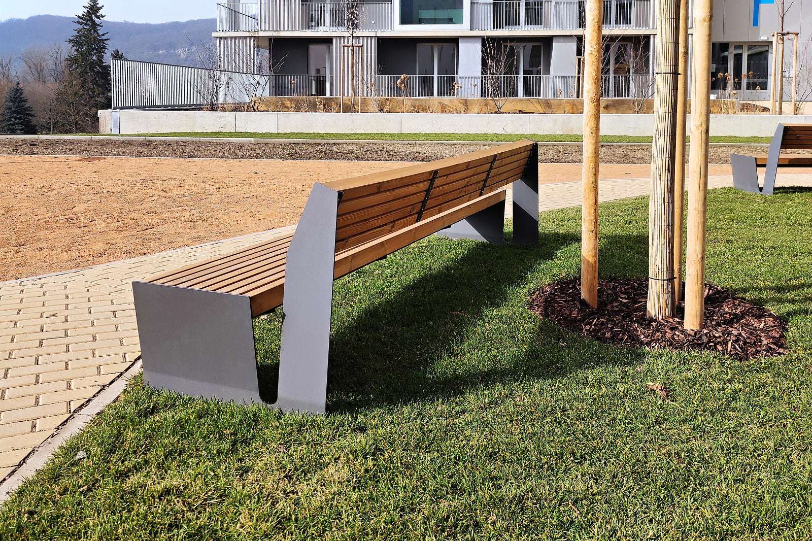 Moderne Holzbank auf Rasen neben gepflastertem Gehweg vor einem Gebäude im Freien platziert.
