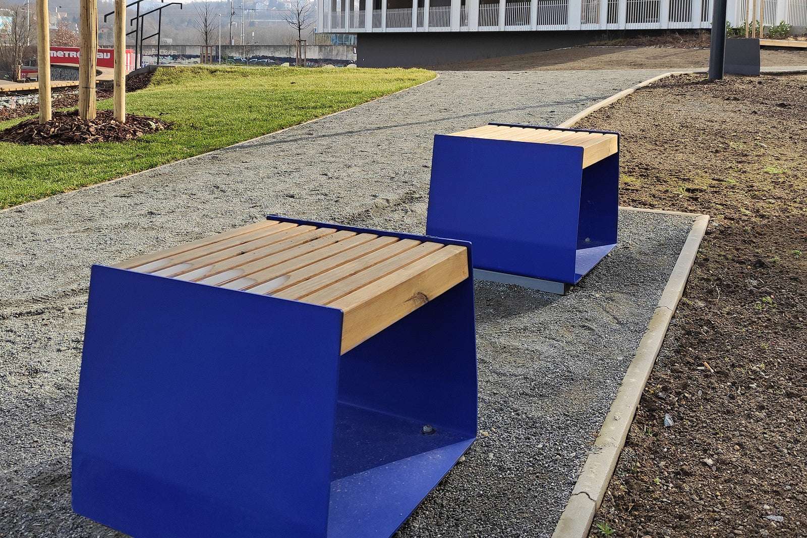 Moderne Bänke mit blauen Seiten und Holzsitzflächen auf einem Kiesweg in einem Gartenbereich.