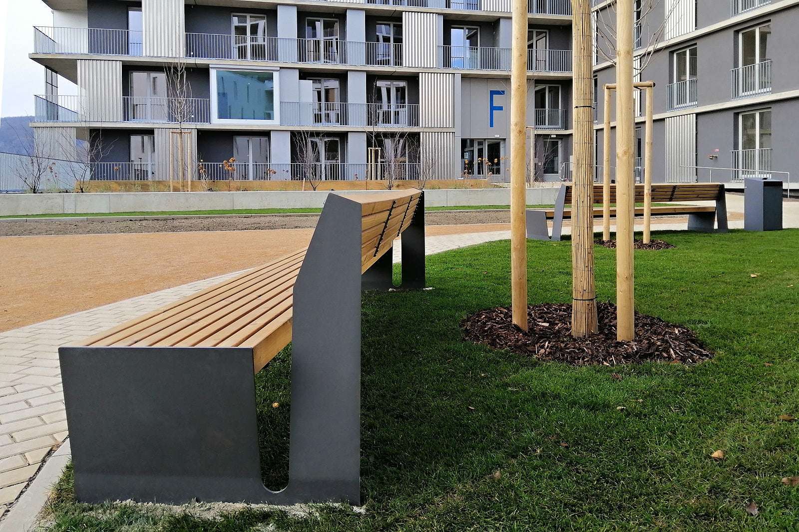 Moderne Holzbänke mit Metallrahmen auf einem begrünten Platz vor einem Wohngebäude.